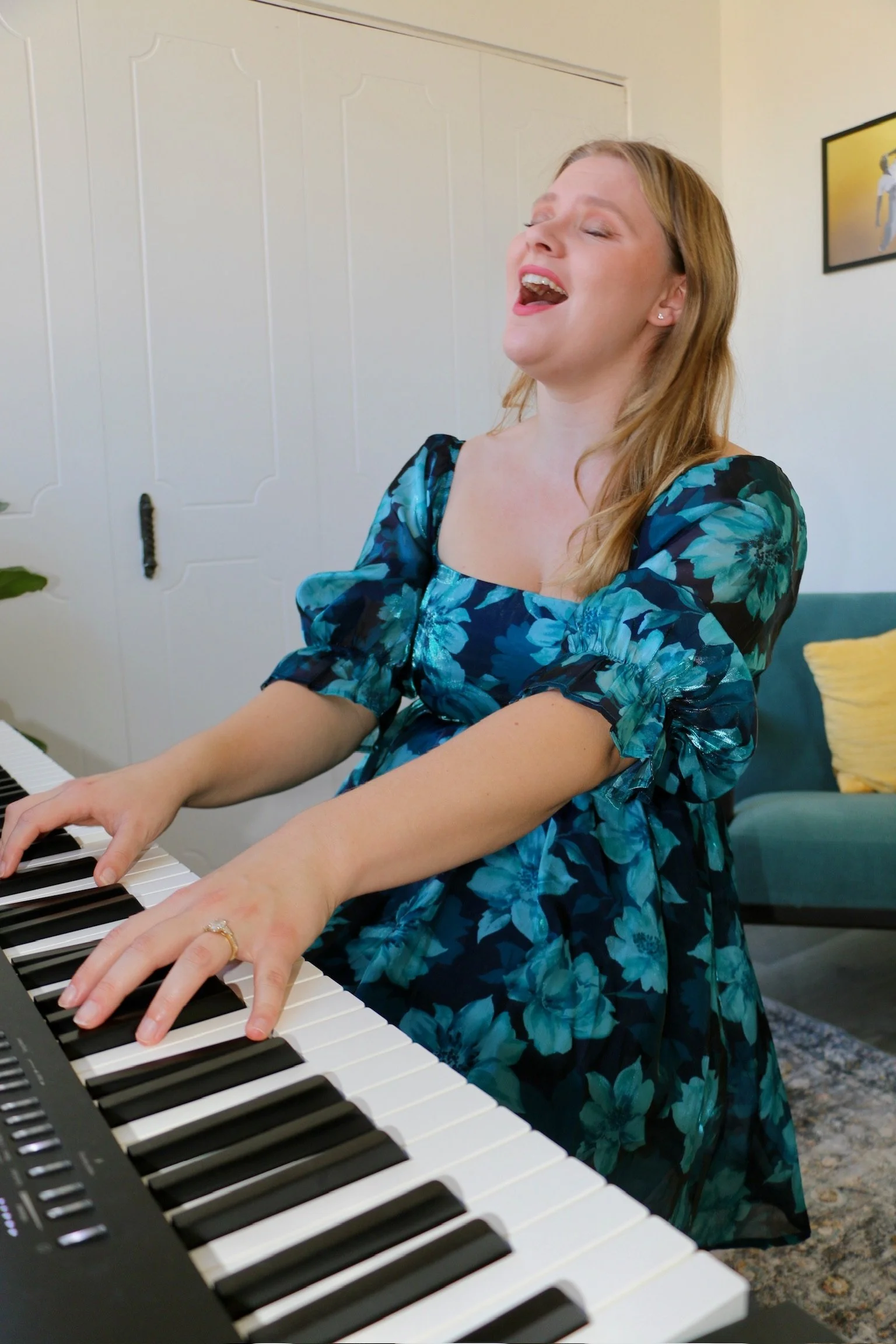 A woman singing and playing the piano indoors, wearing a blue floral dress with short puffy sleeves.