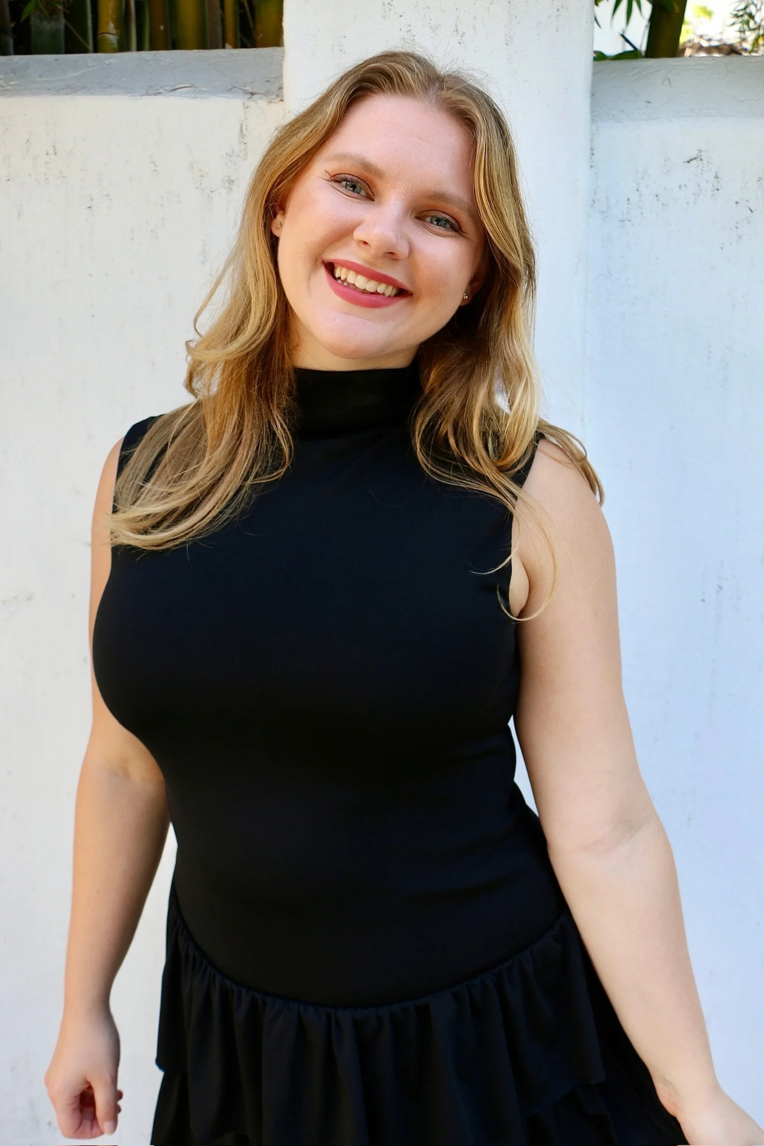 A woman with shoulder-length blonde hair, smiling, wearing a sleeveless black top and a black ruffled skirt, standing outdoors against a white wall with some greenery visible above.