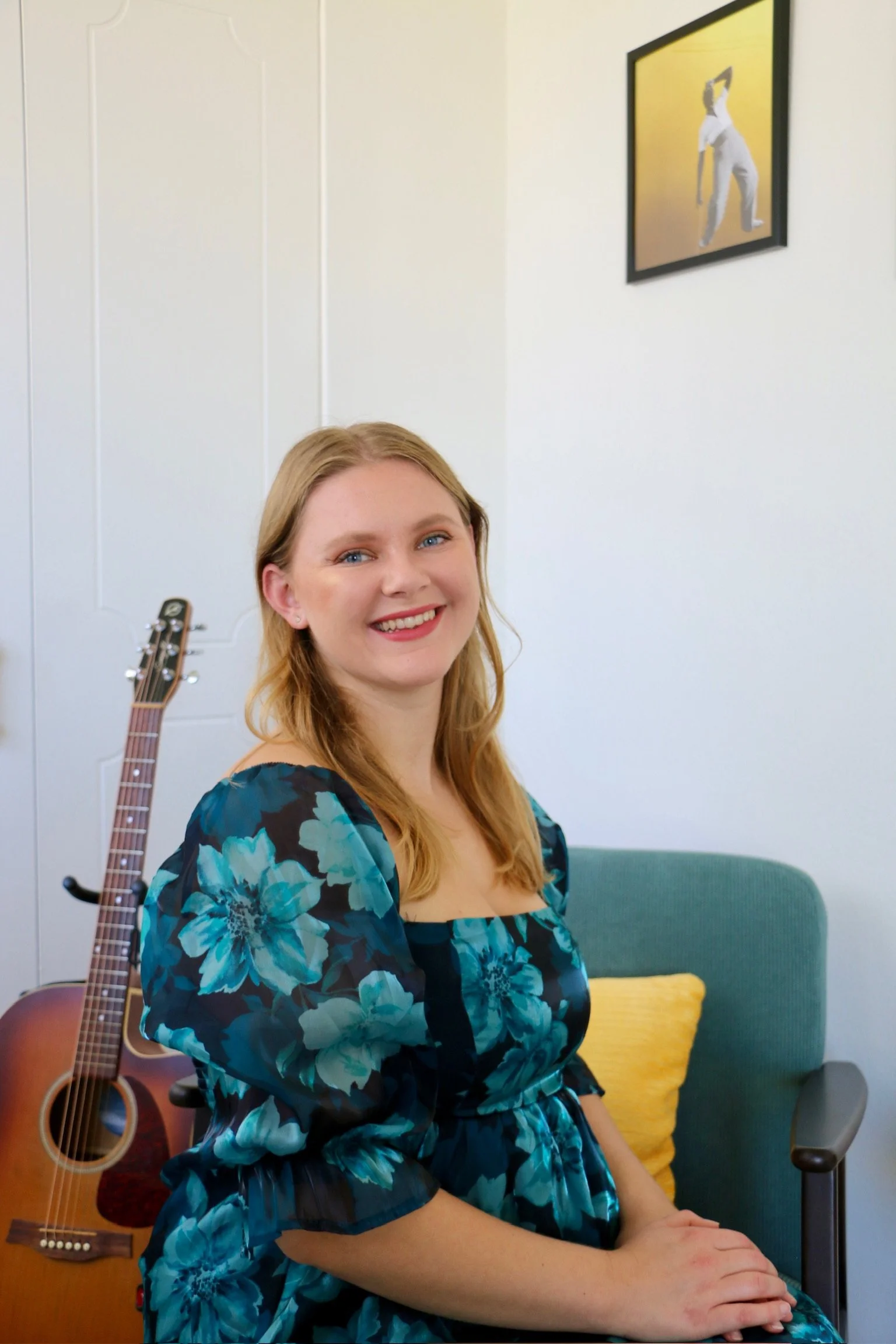 A woman with long red hair smiling, wearing a blue floral dress, sitting on a green armchair in a room with a guitar resting on a stand behind her and a framed picture on the wall depicting a person in white attire performing a dance or gymnastic move.