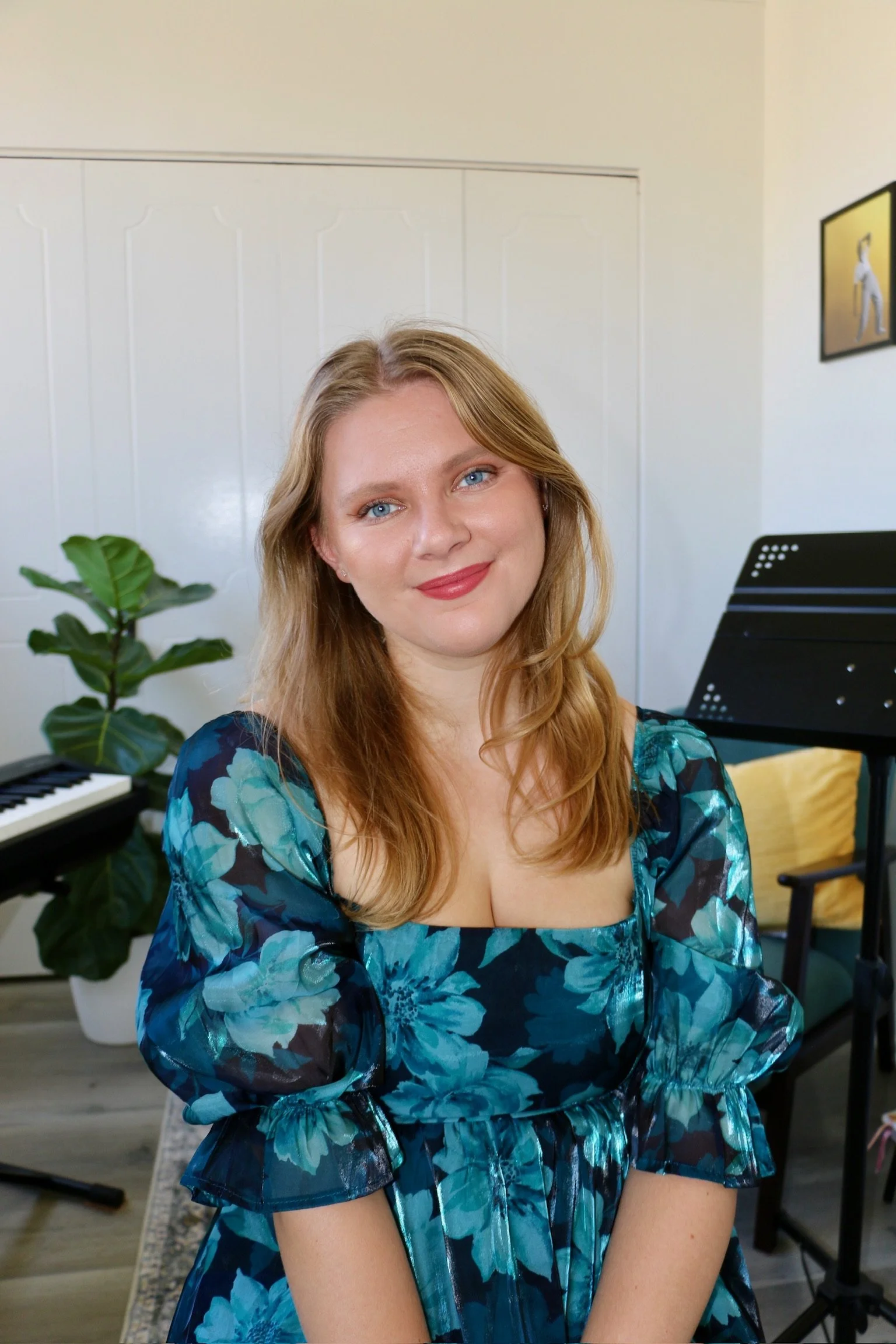 A young woman with long blonde hair and blue eyes, wearing a blue floral dress, sitting in a room with a piano and a music stand, smiling at the camera.