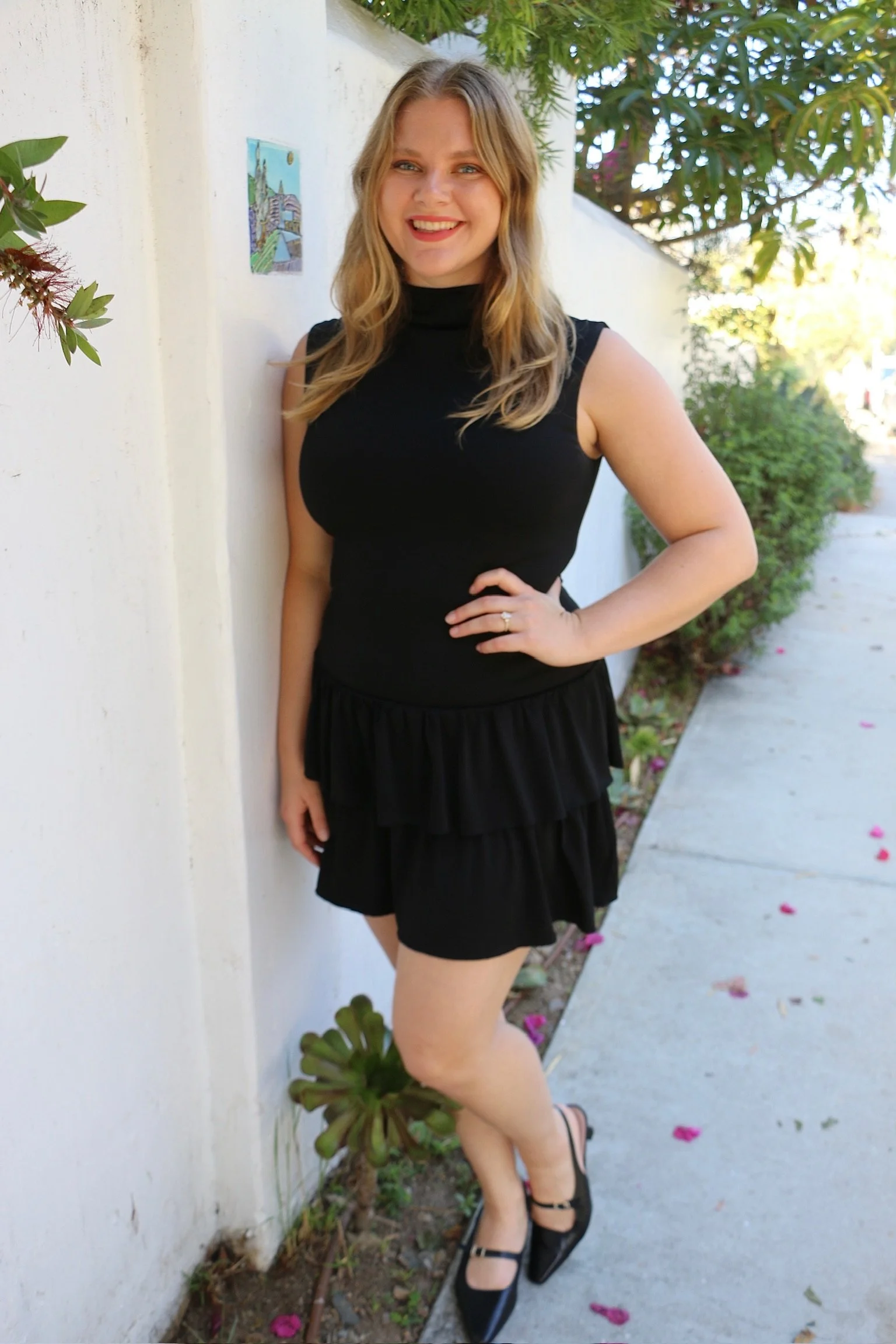 A woman with blonde hair and a black sleeveless dress standing outdoors, leaning against a white wall, smiling, with green foliage and a sidewalk in the background.