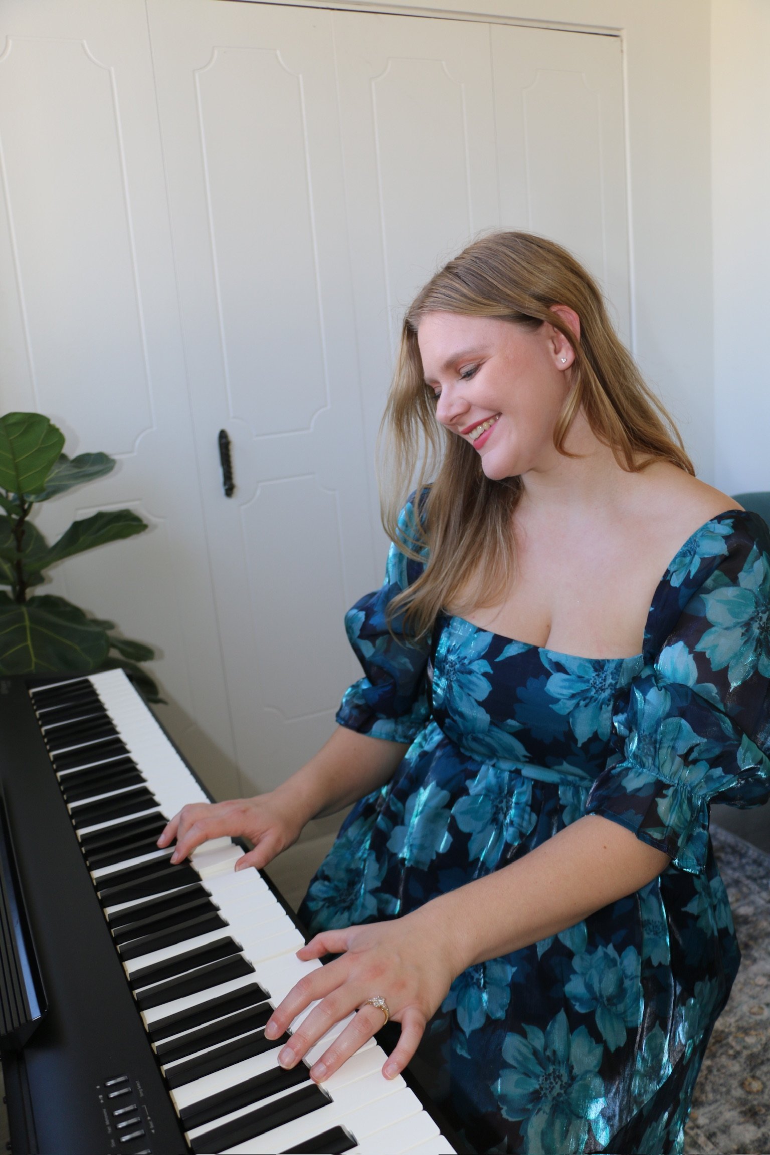 A woman with long, light brown hair is smiling while playing a black digital piano. She is wearing a blue floral dress with puffed sleeves and a ring on her left hand. There is a large leafy houseplant to her left and a white door behind her.