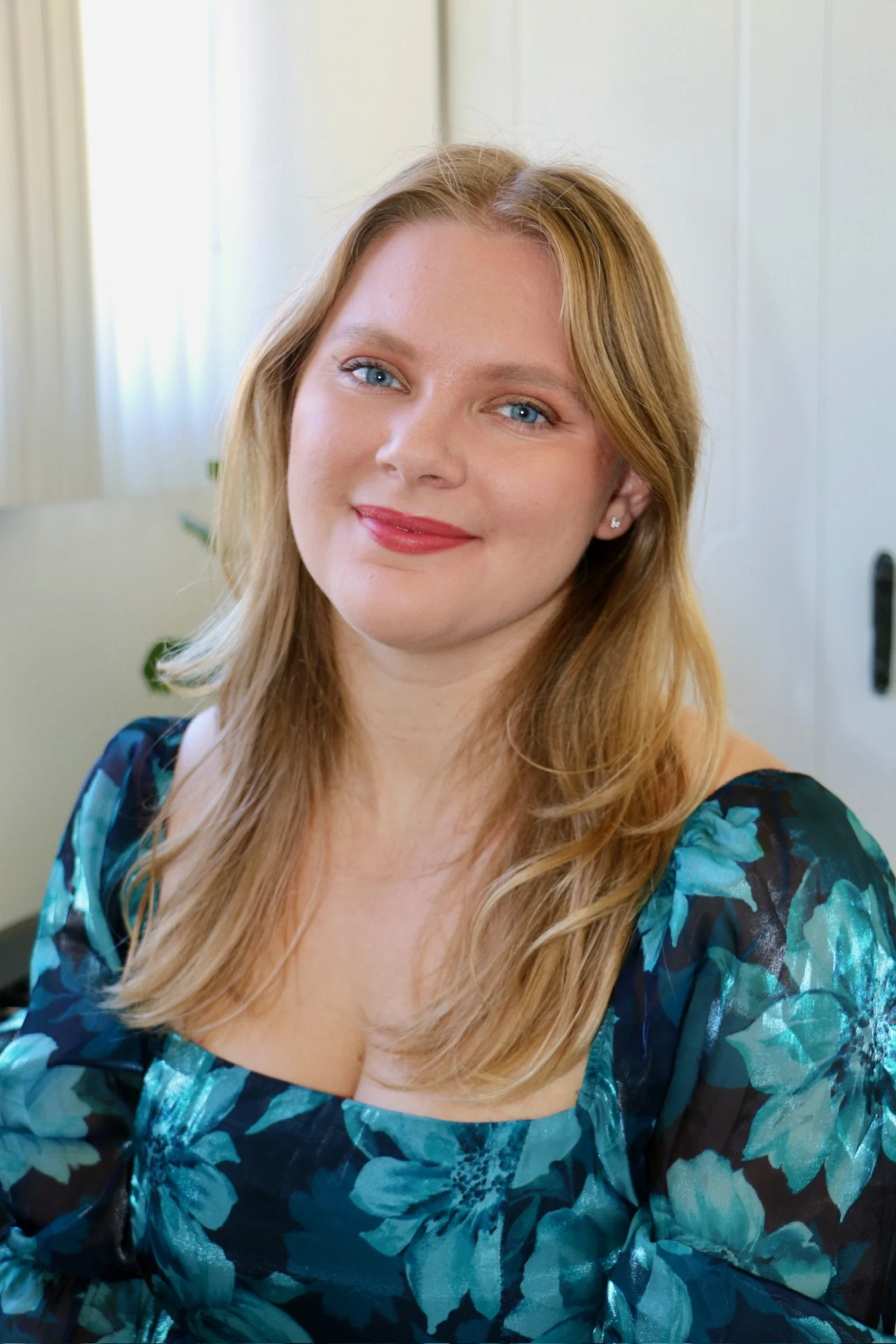 A woman with long red hair and blue eyes smiling, wearing a blue floral top, in a room with white walls and a closed door.