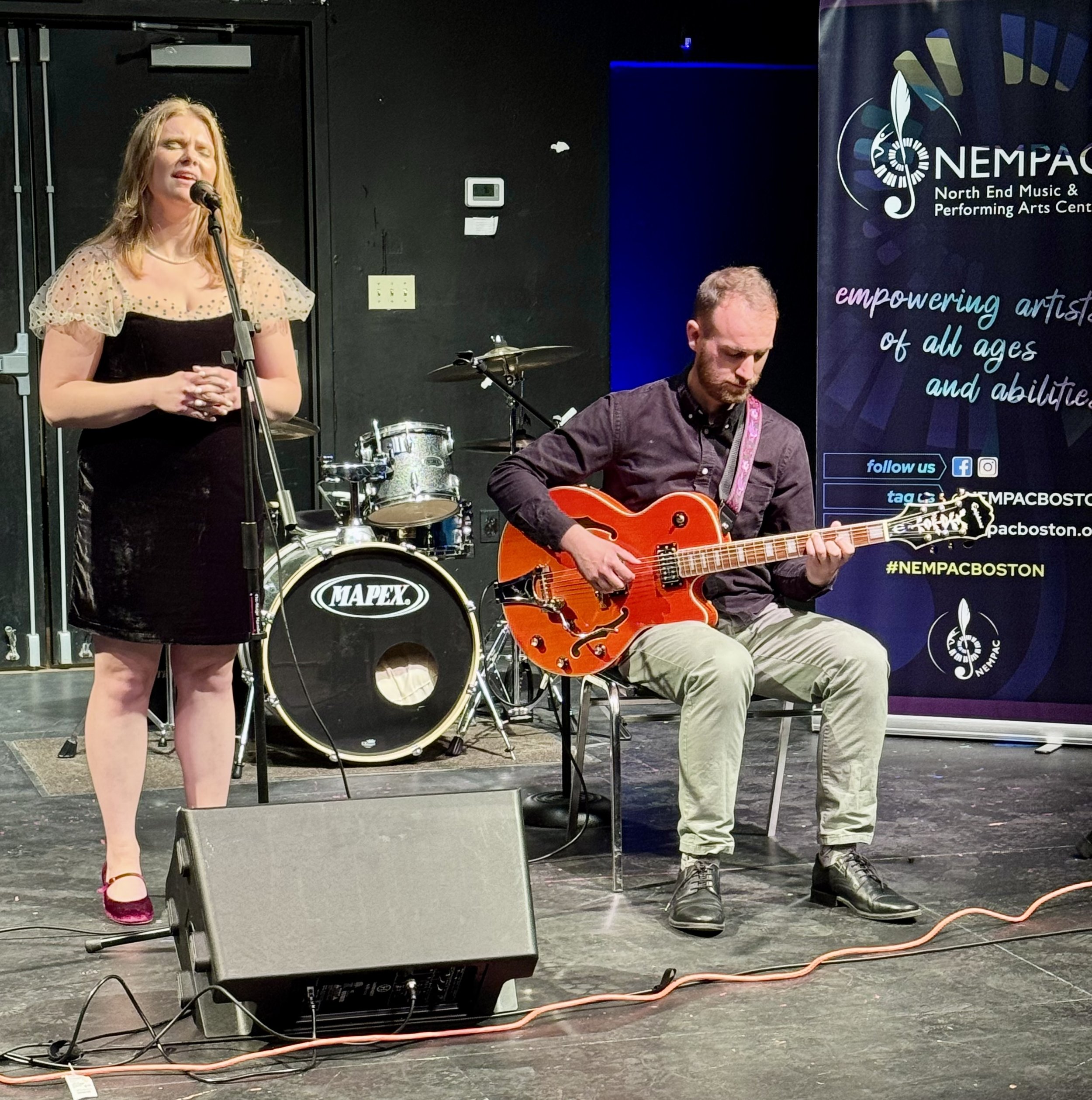 A woman singing into a microphone on stage, and a man playing an orange electric guitar, with a drum set behind them and a banner for NEMPAC Boston in the background.