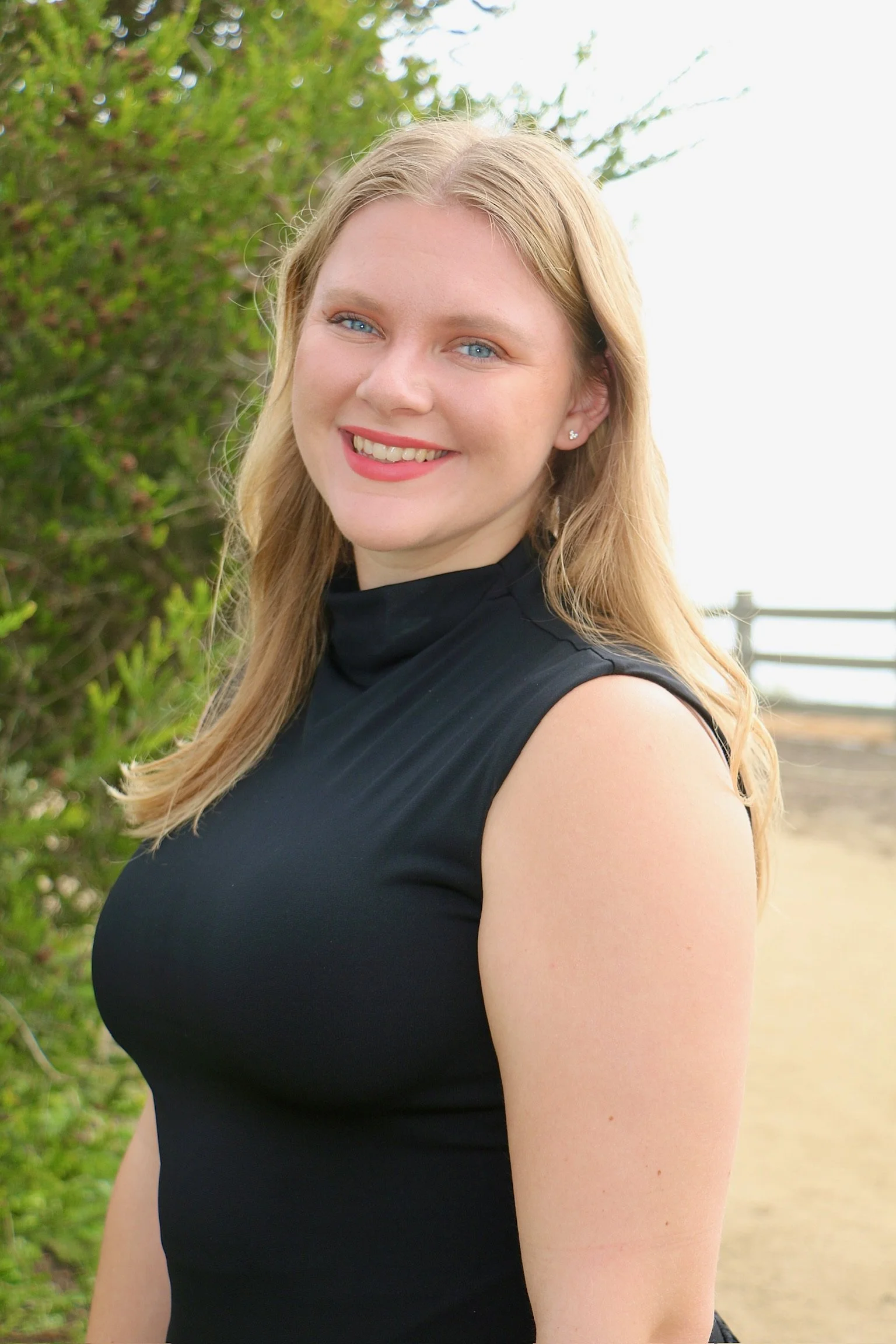 A young woman with blonde hair, blue eyes, and a bright smile, standing outdoors with greenery and a fence in the background.