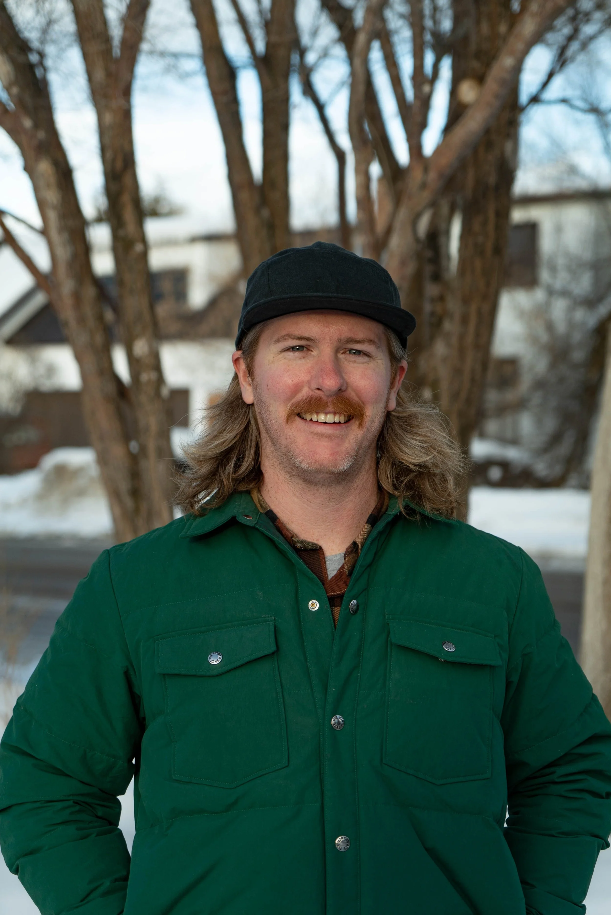 Smiling man with long hair, mustache, and beard, wearing a black cap and green jacket, outdoors during winter.
