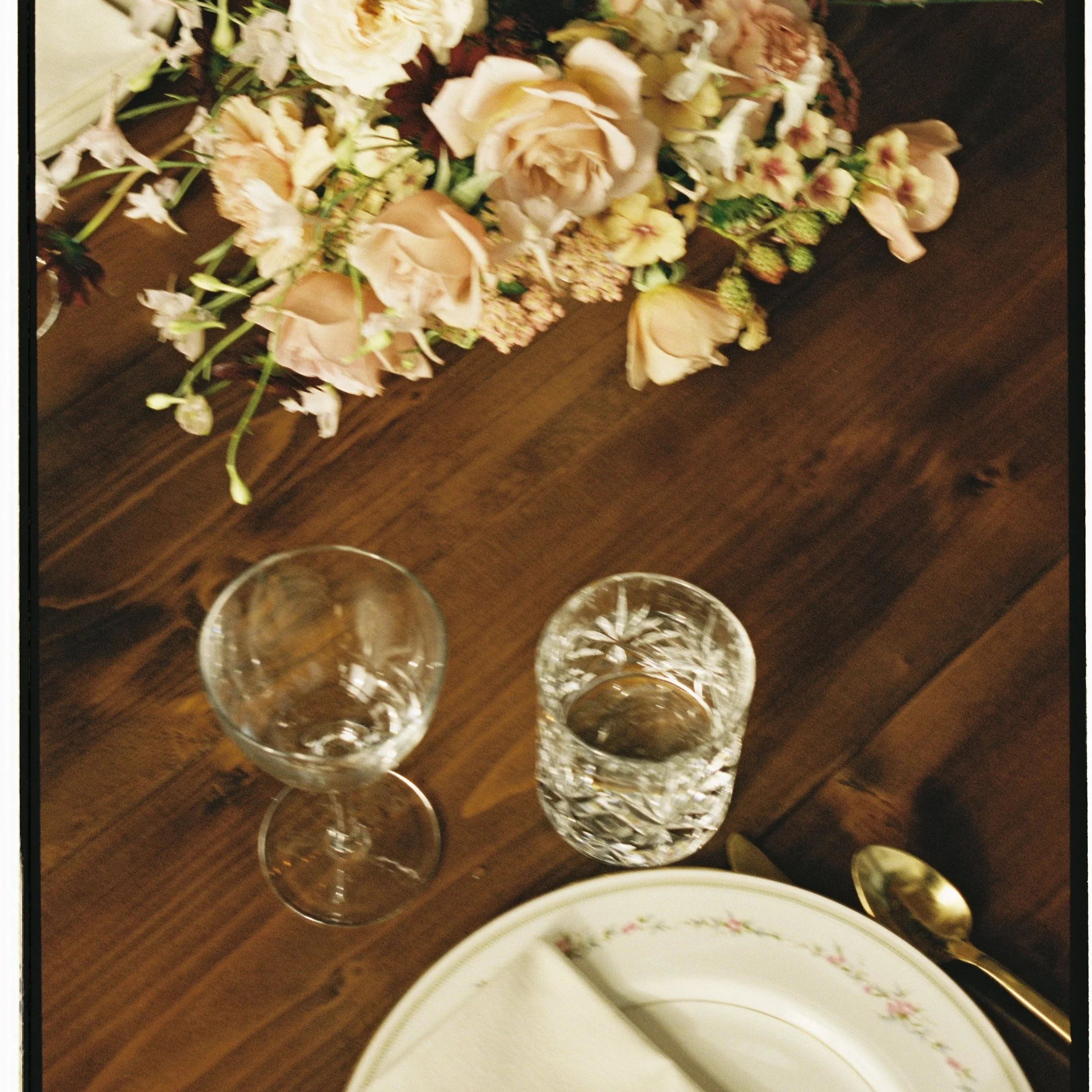 A table setting with an ornate plate, silverware, a napkin, two empty glasses, and a floral centerpiece with pink and white flowers on a wooden table.