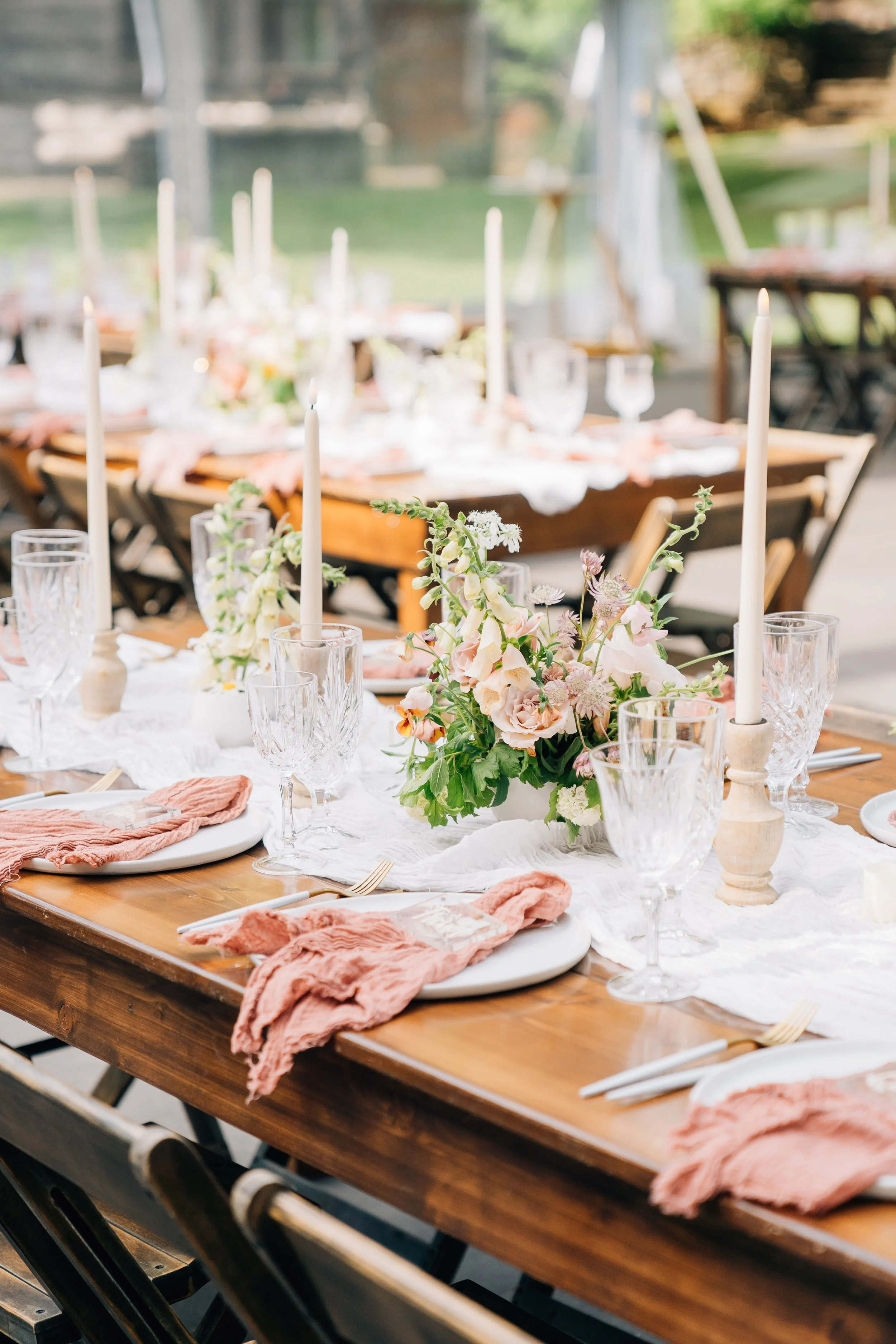 Decorated outdoor dining table with pink napkins, clear glassware, a white runner, pink and white floral centerpiece, and tall white candles.