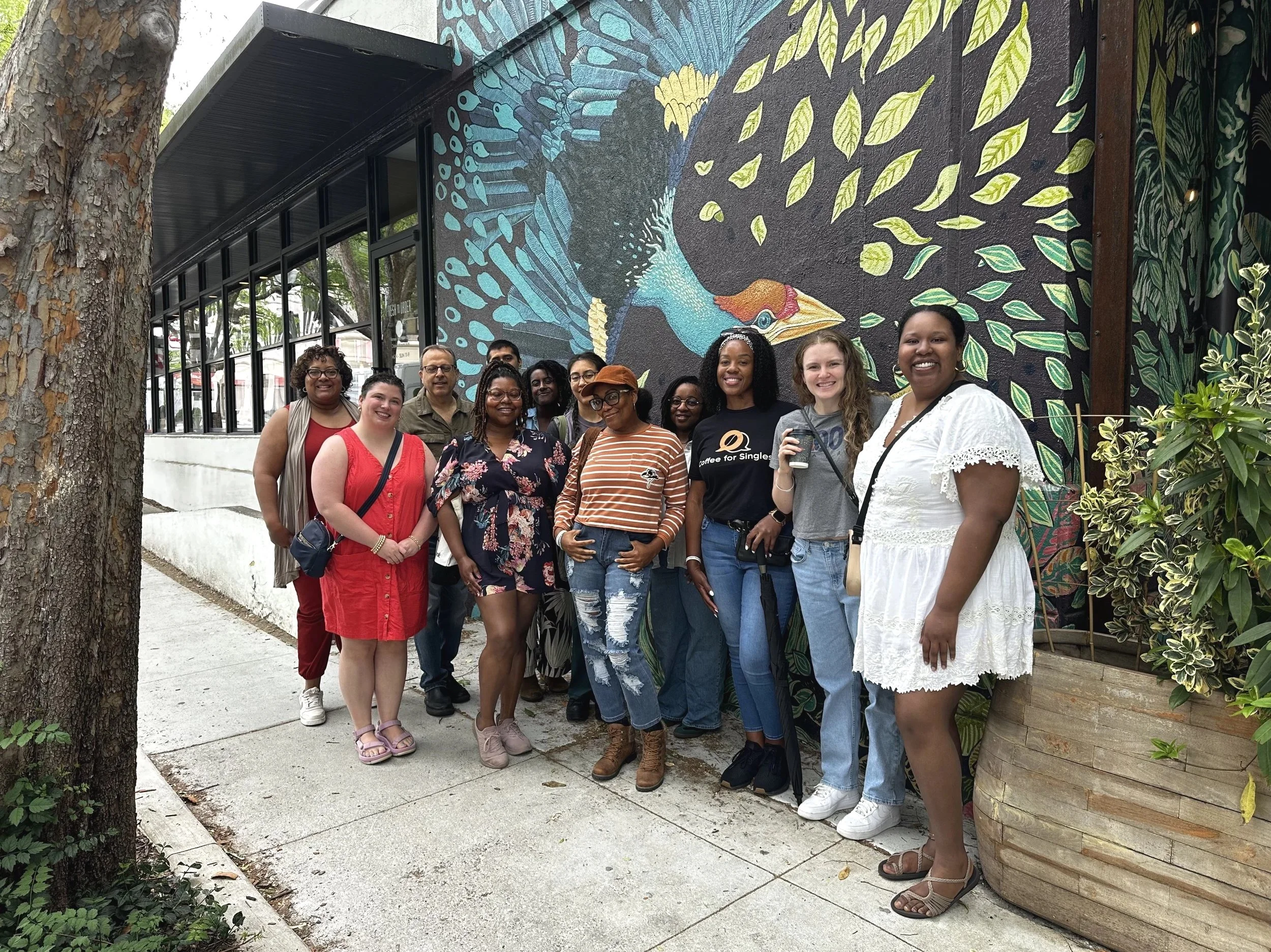 A group of diverse people standing outdoors in front of a colorful mural of a peacock with vibrant blue feathers and green leaves. They are smiling and posing for the photo.