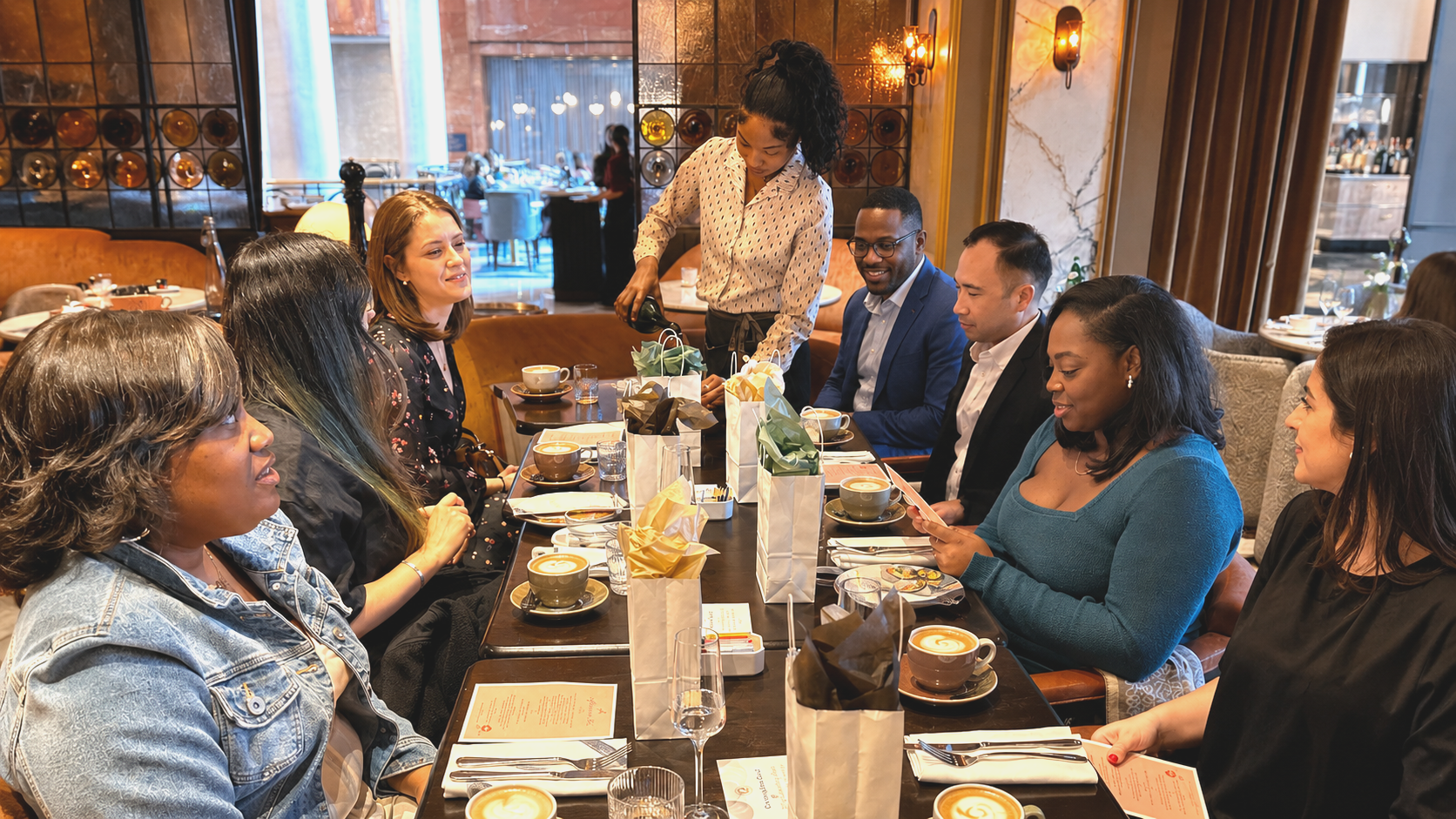 A group of diverse people sitting around a dinner table in a restaurant, with a woman serving coffee and others engaged in conversation.