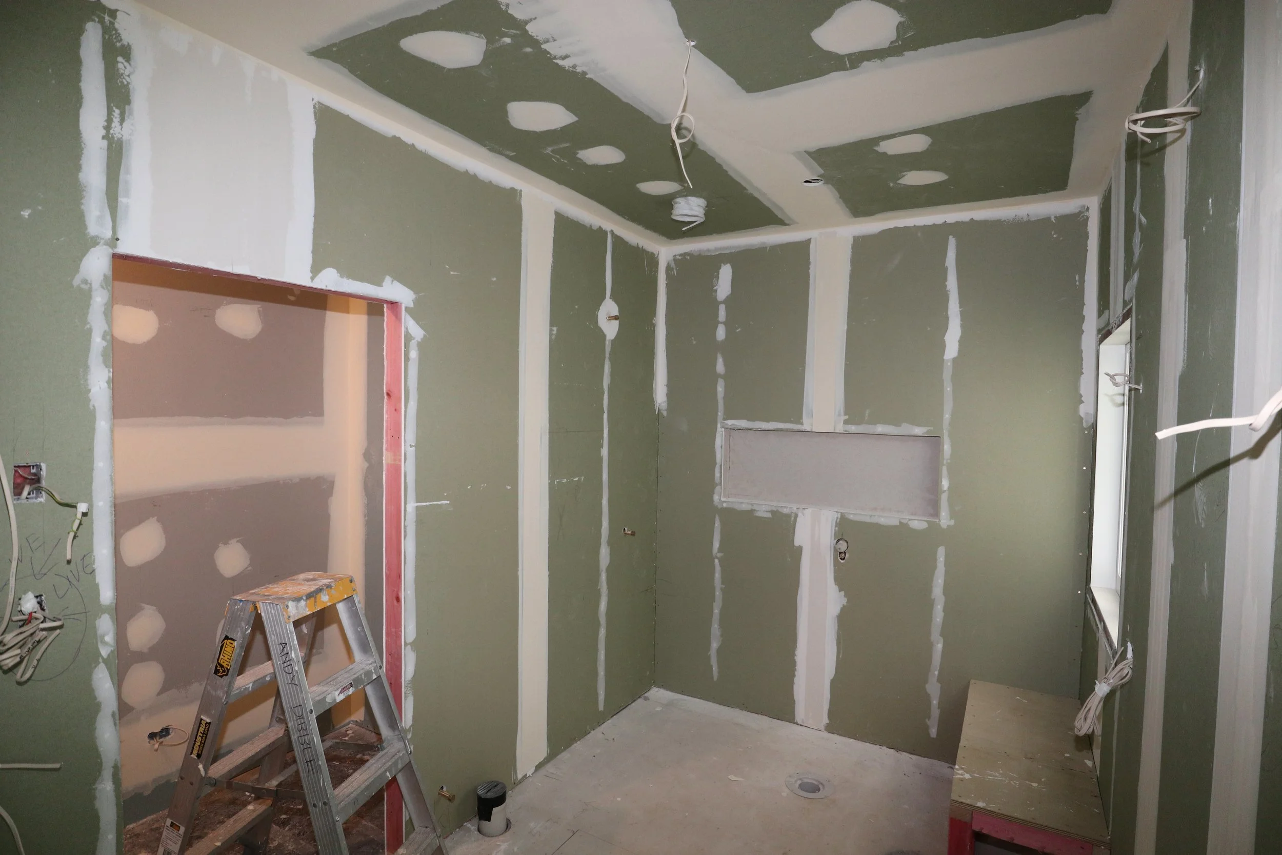 Bathroom under renovation with green drywall and white drywall tape, wiring hanging from ceiling, small window, ladder, and plumbing fixtures.