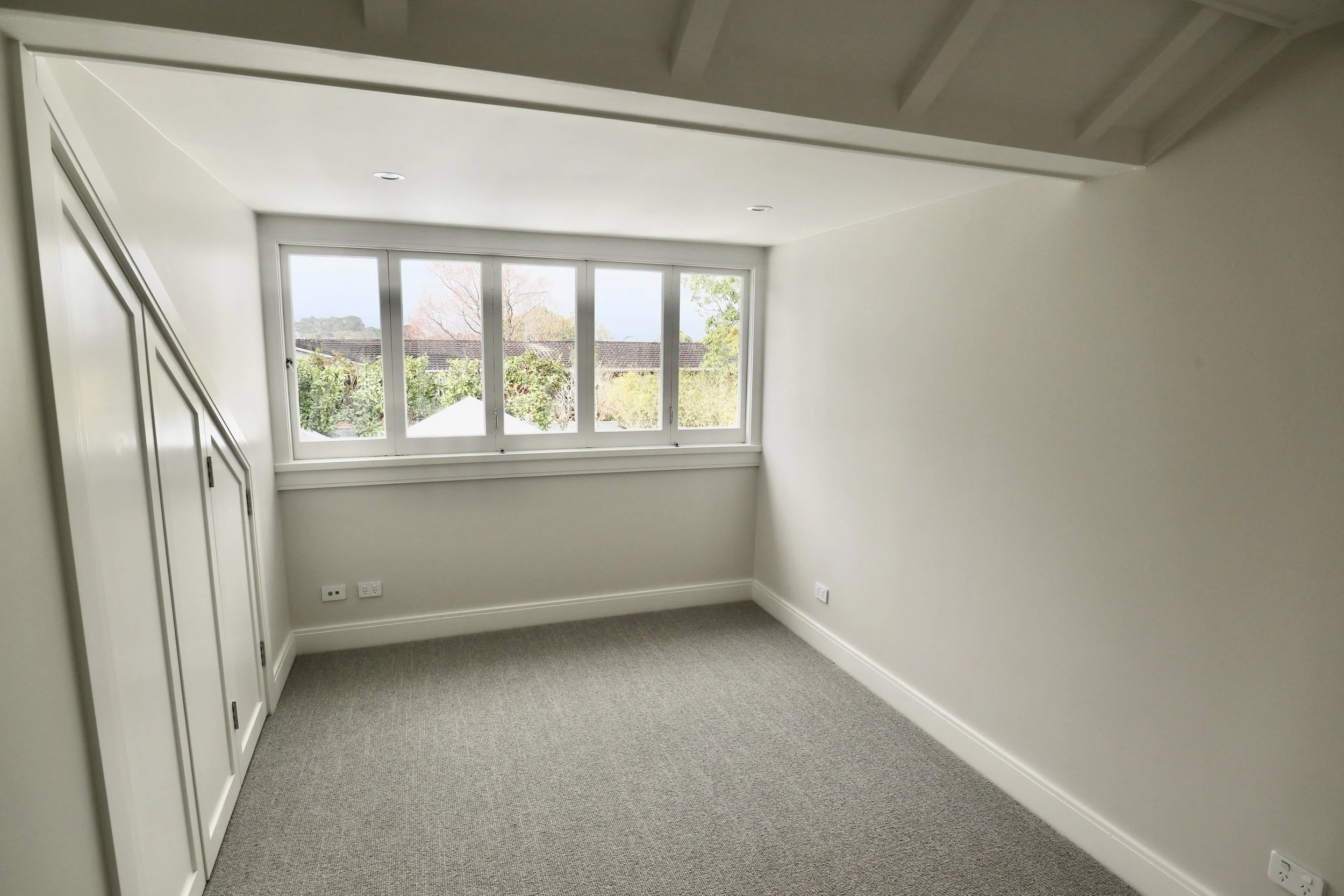 Empty room with large window showing trees and sky outside, white walls, gray carpet, built-in white closet, and electrical outlets.