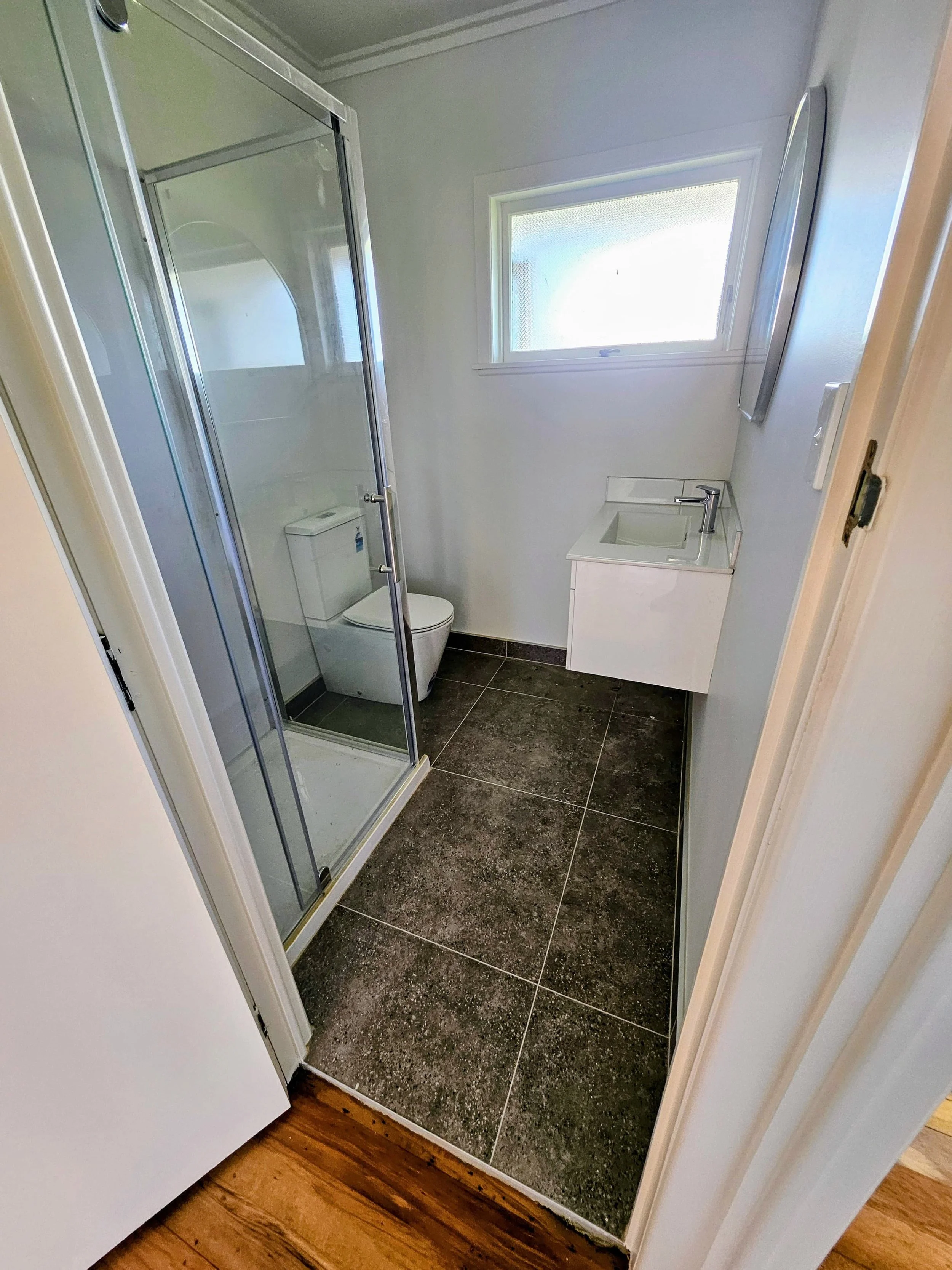 Small bathroom with a glass shower stall, a toilet, a small white sink with a cabinet, and a window above the sink.