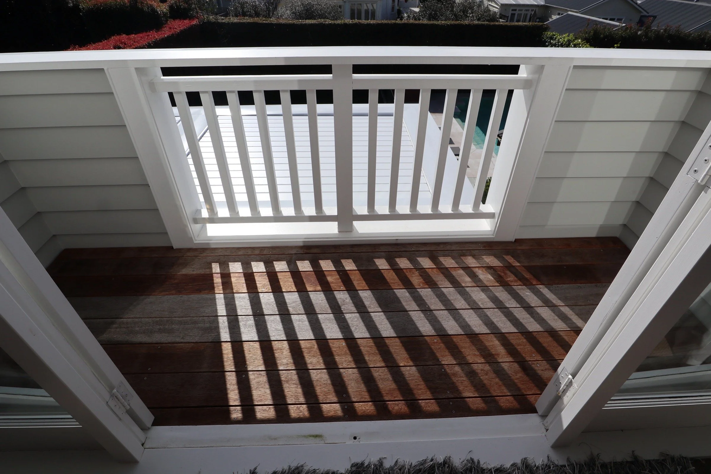 Small balcony with wooden floor, white railing with vertical slats, and sliding glass door leading inside. Shadows from the railing create striped patterns on the floor.