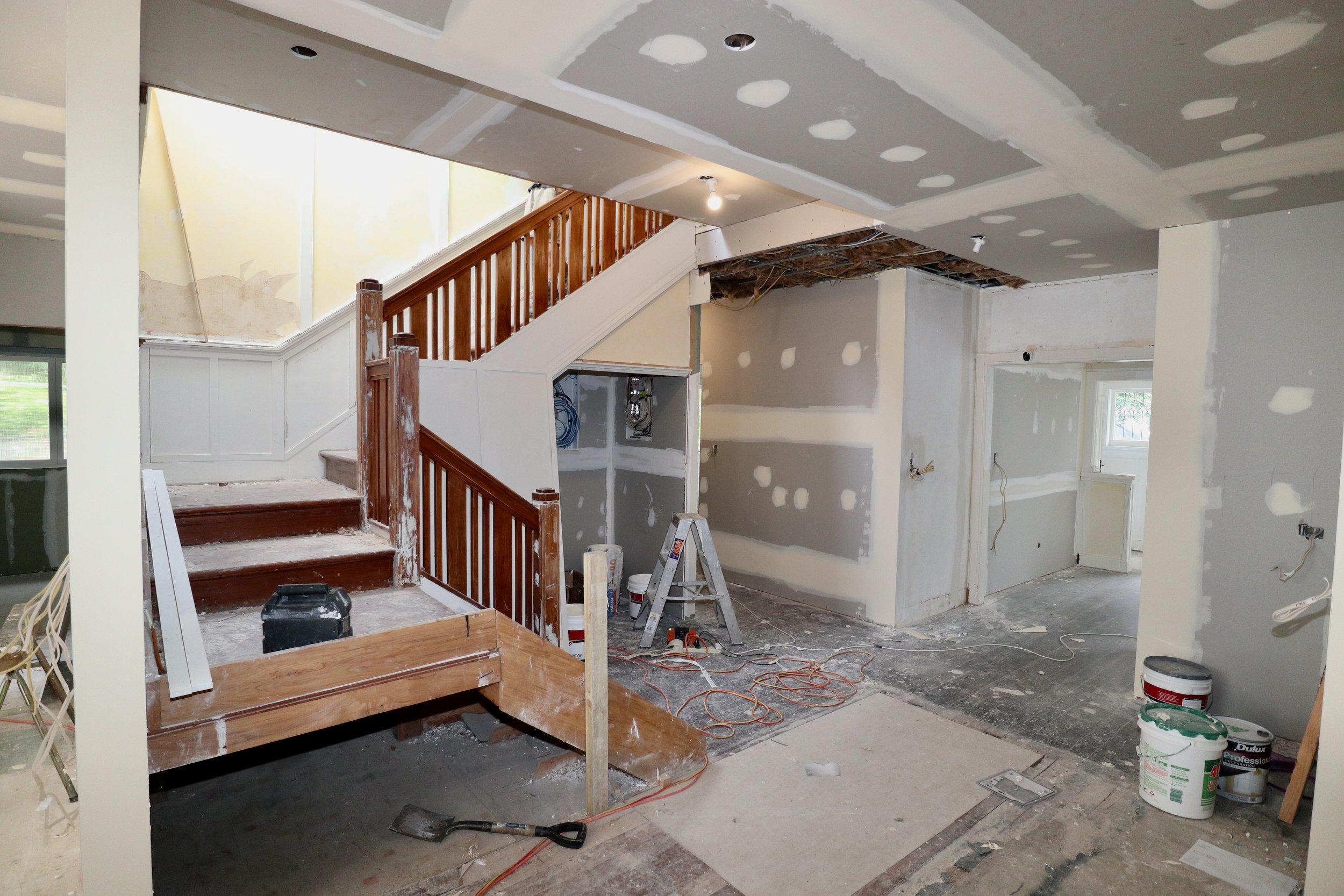 Interior of a house under renovation with unfinished walls, drywall, and construction tools. A staircase with wooden banisters leads upward, and there are construction materials scattered on the floor.