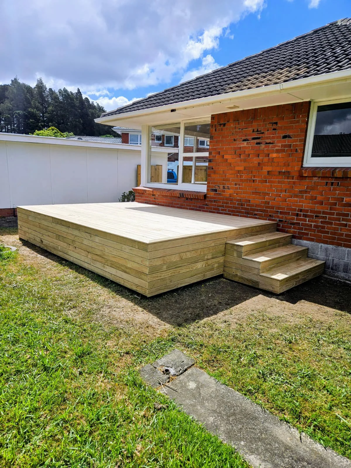 Newly built wooden deck outside brick house with stairs, set on grassy yard under partly cloudy sky.