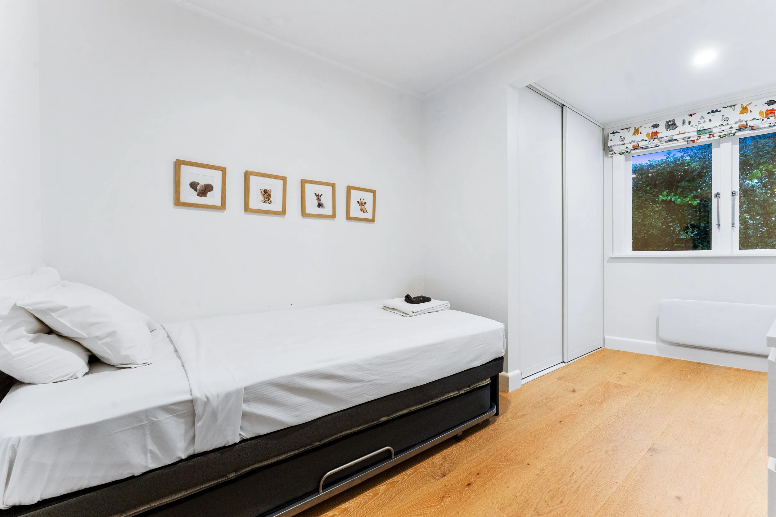 A bedroom with a single bed, white bedding, and pillows, decorated with four framed animal pictures on the wall, and a window with views of trees and a curtain rod with animal-themed curtains.