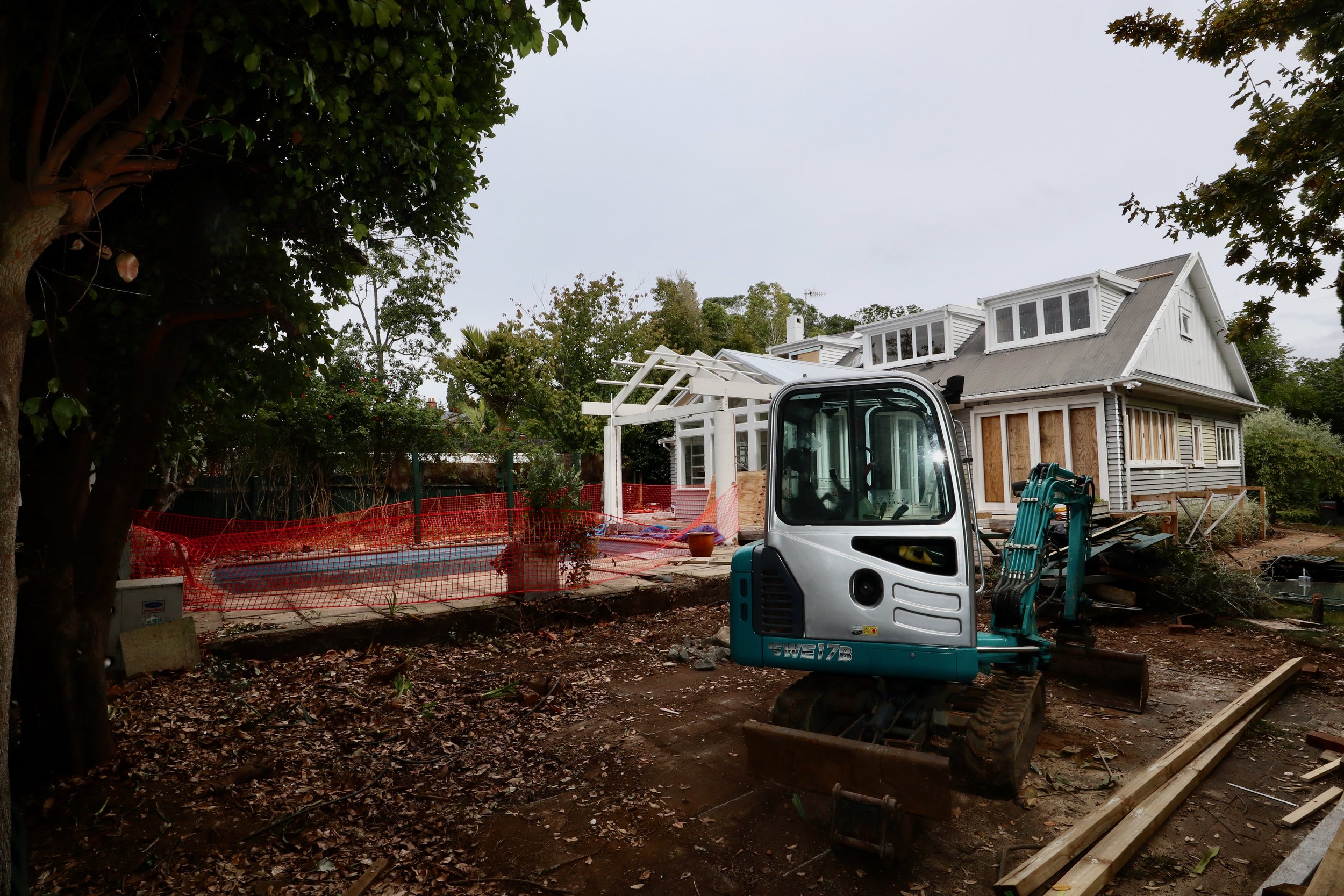 House under renovation with construction equipment and materials in the yard, surrounded by trees and fenced off area.