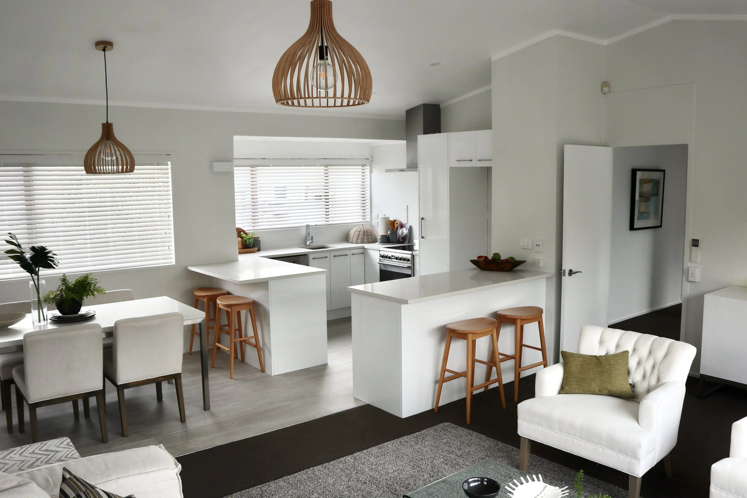 Open-concept living room and kitchen with white walls, gray floors, white cabinetry, and wooden accents including pendant lights and stools. Features a dining table with five chairs, a white armchair with a green pillow, and a kitchen counter with st