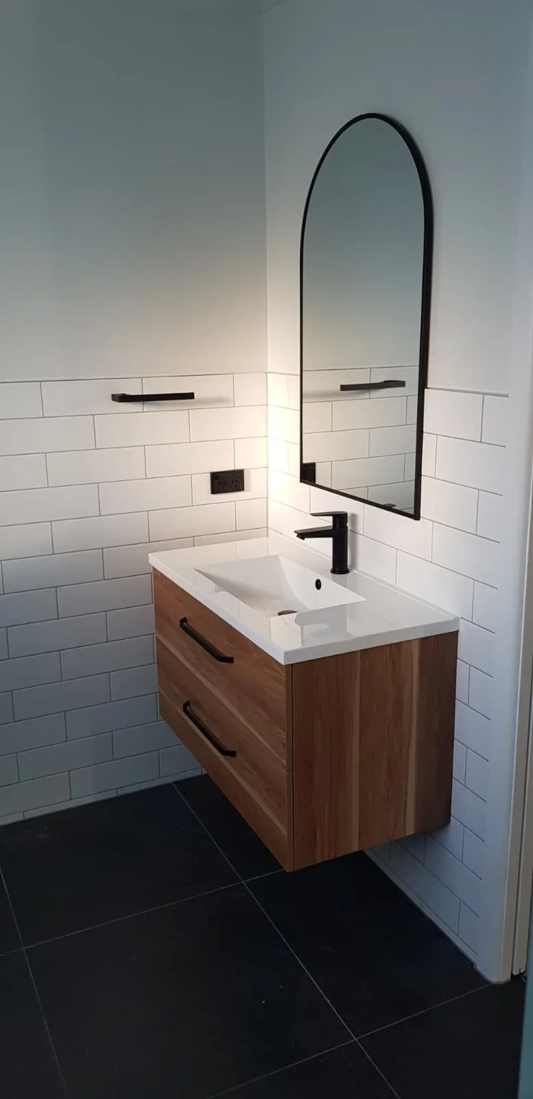Modern bathroom vanity with a white rectangular sink, black faucet, black horizontal handles on the wooden cabinet drawers, a large mirror with a black frame, and white subway tiles on the wall. Black electrical outlets on the wall and black floor ti