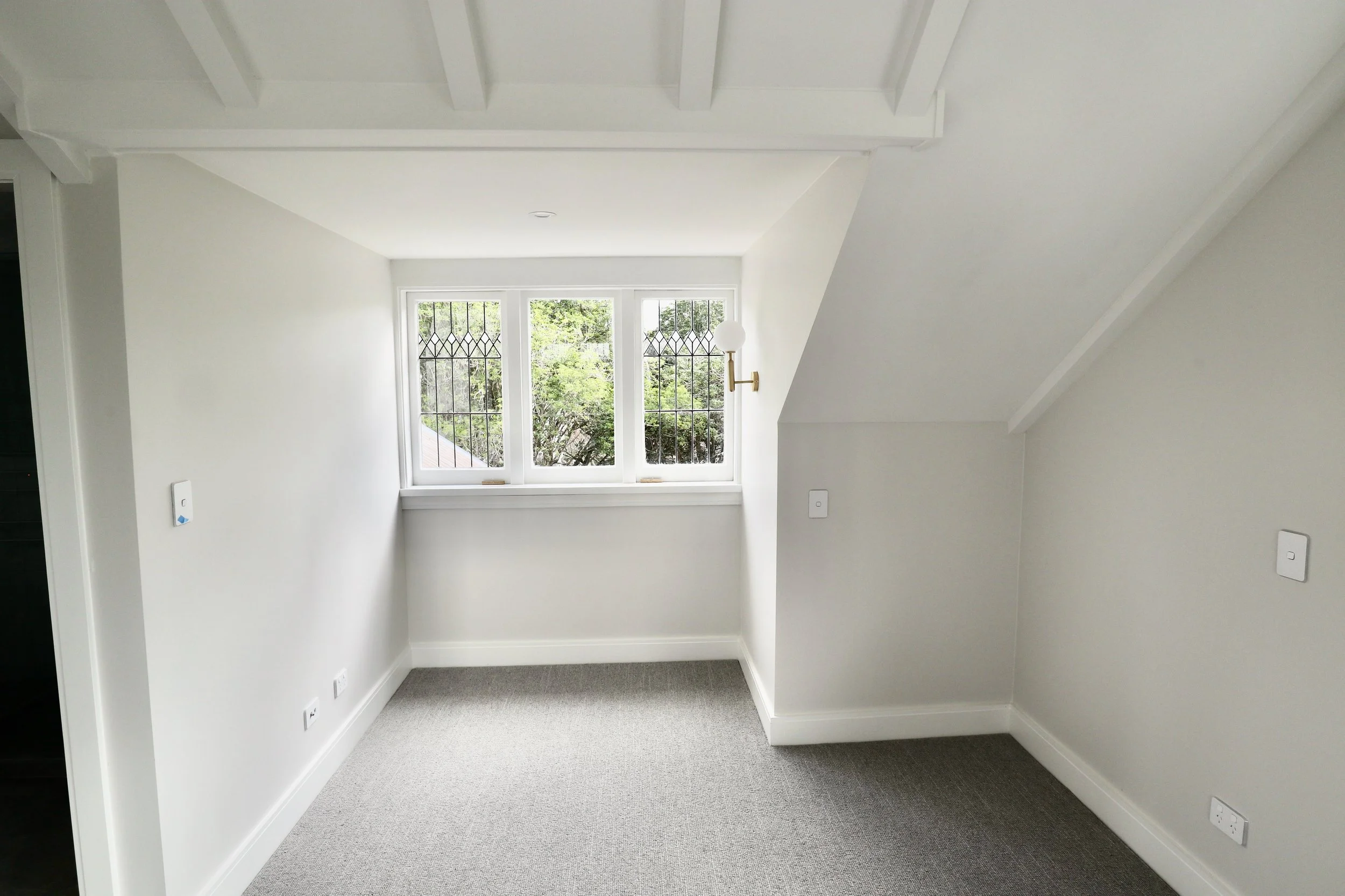 Empty room with white walls, gray carpet, and a window with a view of green trees, featuring a brass wall sconce with a white globe light.