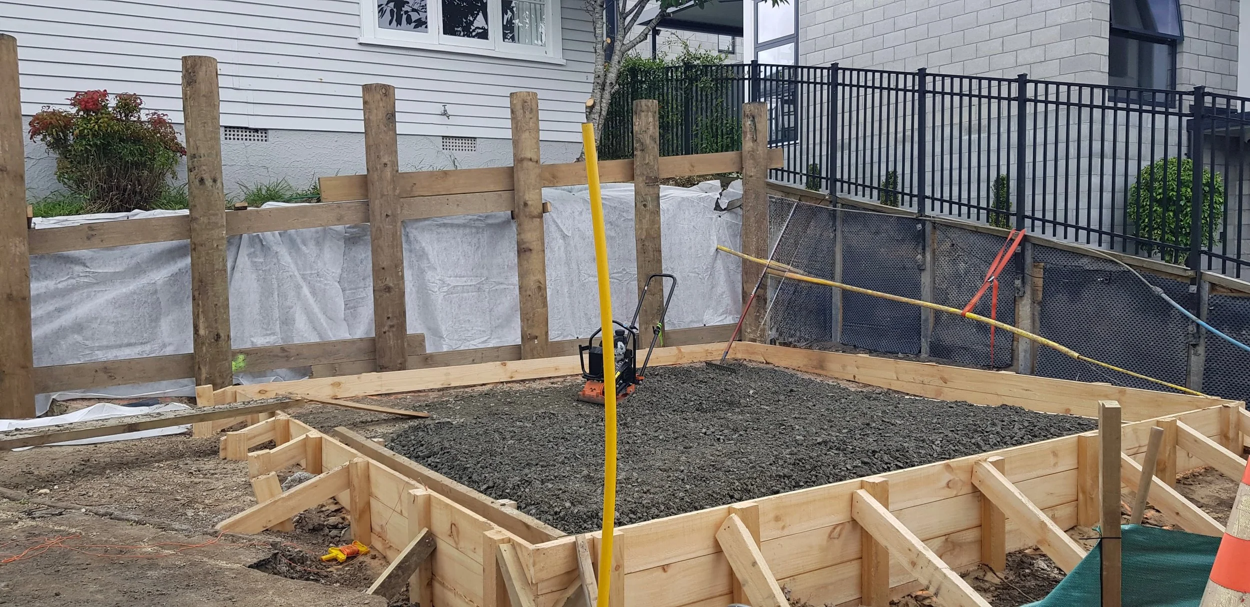 Construction site showing a wooden foundation with gravel, surrounded by a retaining wall made of wood and reinforced with nails, in a residential area with houses and trees in the background.