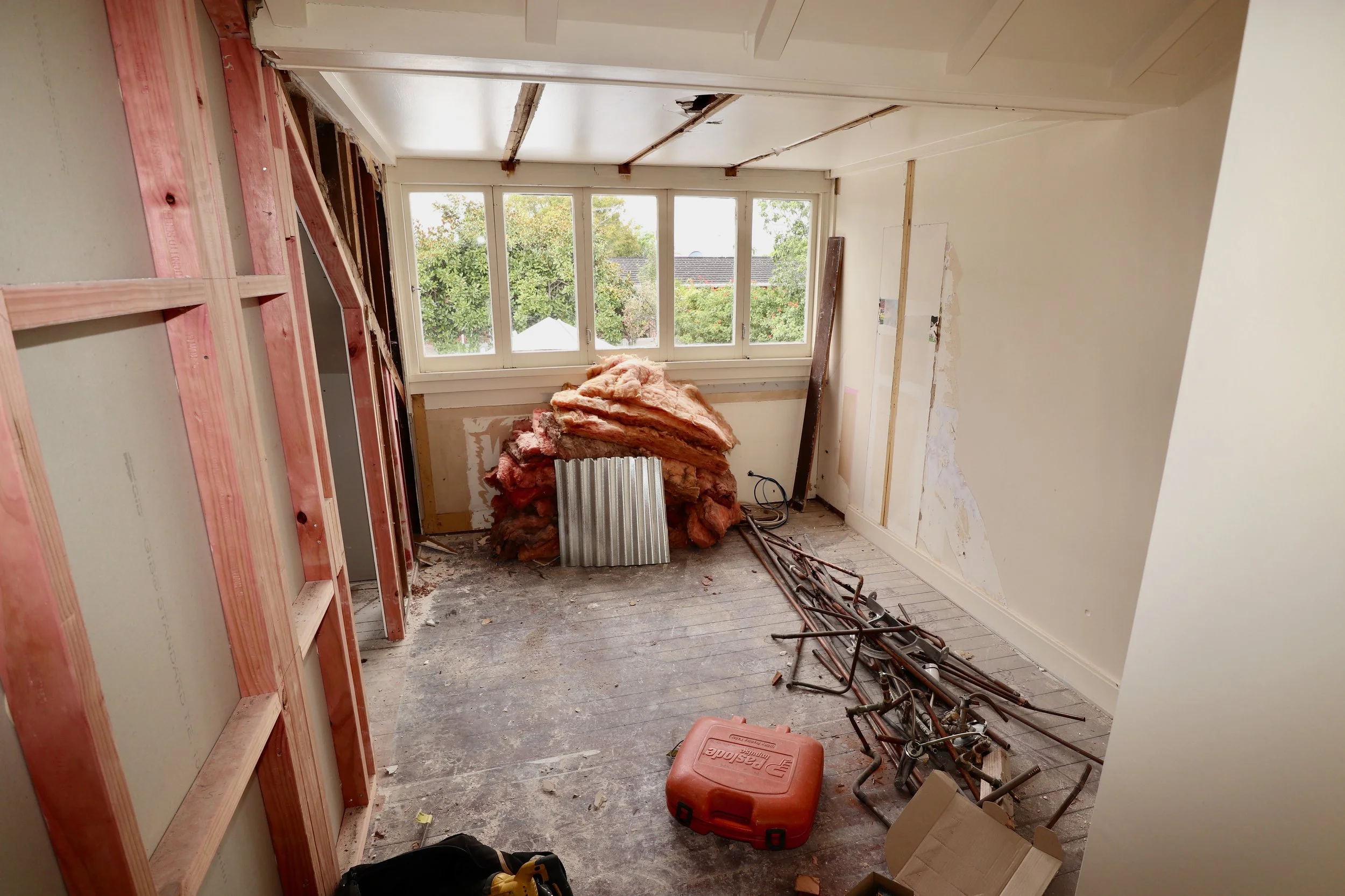 Room under renovation with exposed insulation, construction tools, and partially removed walls, large window at the back showing trees outside.