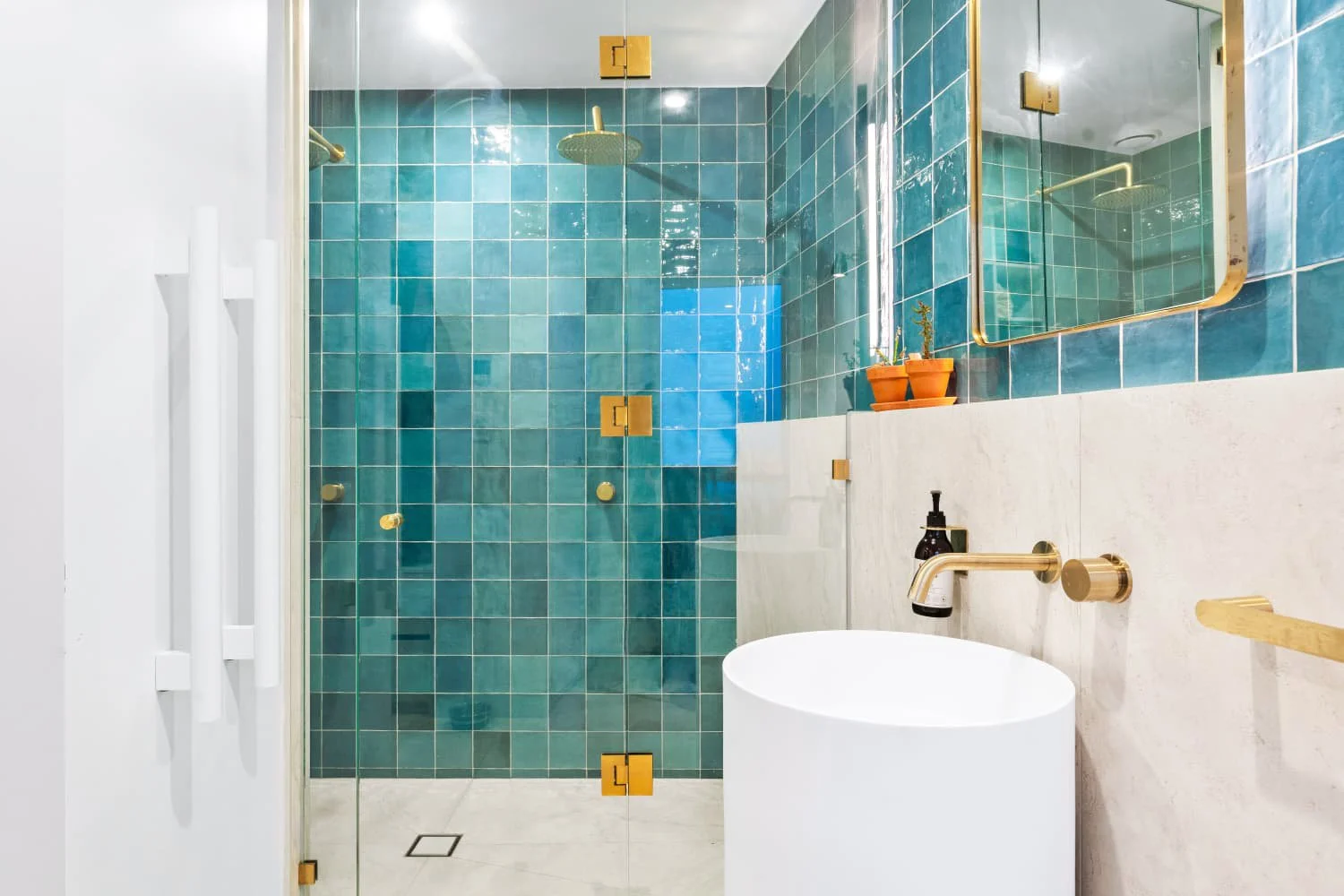 Modern bathroom with teal tiled shower area, gold fixtures, a white rounded sink, and a large mirror.