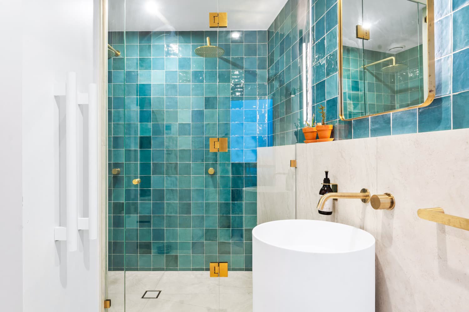 Modern bathroom shower with teal tiles, glass door with gold hardware, white freestanding sink, gold faucet, mirror, and small potted plants on a shelf