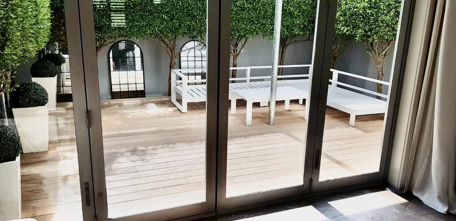 A view of a wooden deck outside through glass sliding doors, with benches, potted topiary, and a small tree against a wall with arched windows.