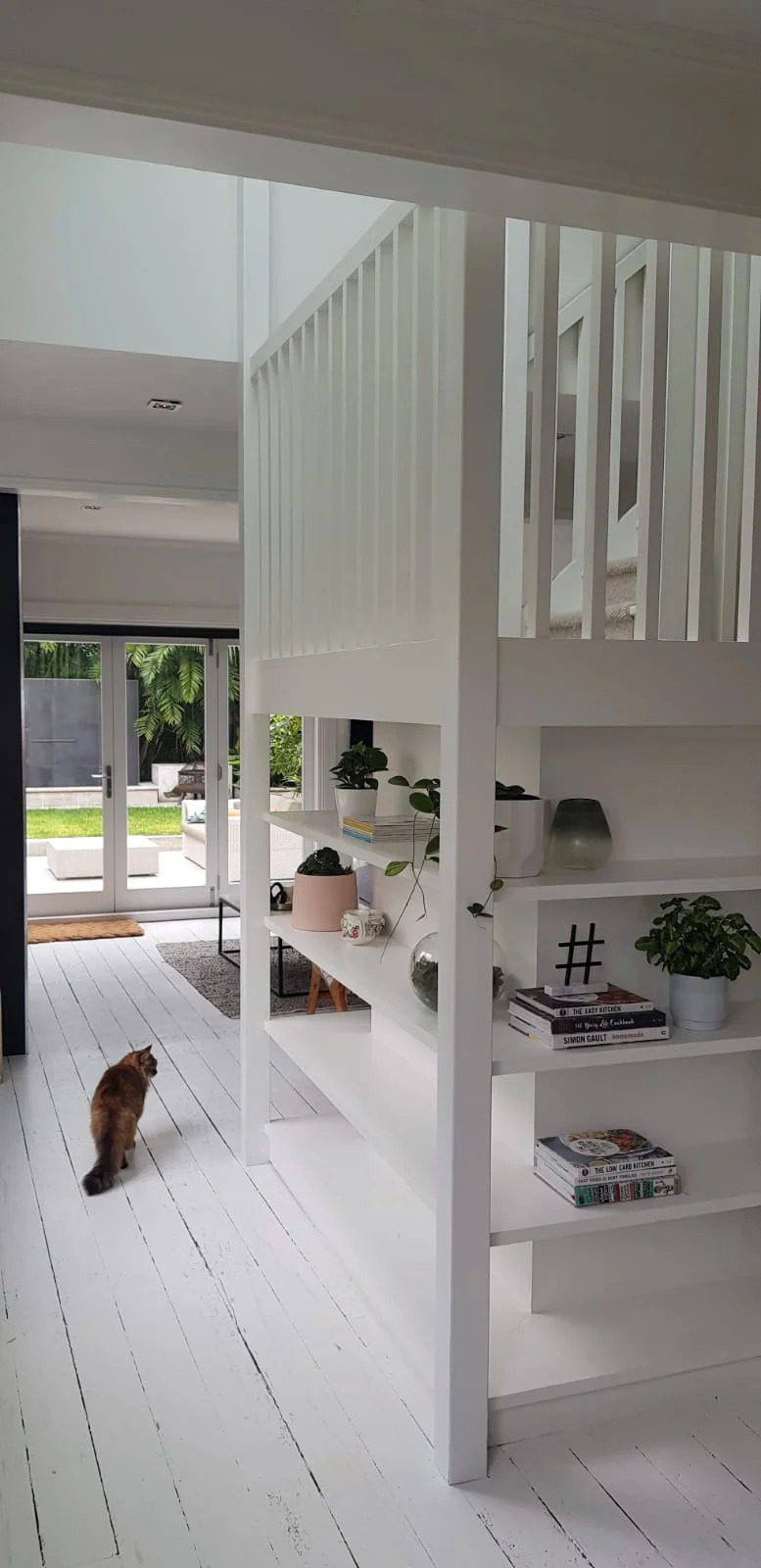 Interior of a modern home with a white wooden floor, white stairs with vertical railings, and a built-in white bookshelf. There is a cat walking on the floor near the bookshelf, and glass doors leading to a backyard with greenery and outdoor furnitur