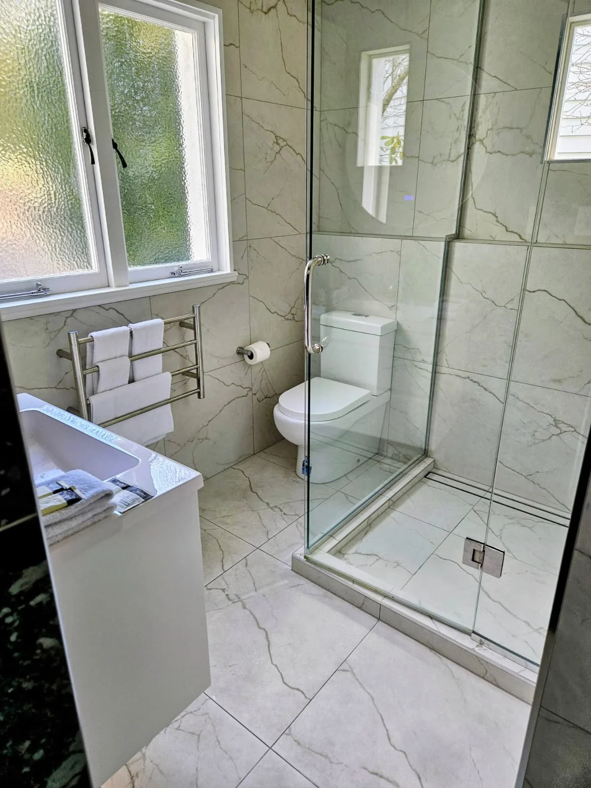 Modern bathroom with large marble tiles on the walls and floor, a frosted window, a white toilet, a glass shower enclosure, a towel rack with two white towels, and a small sink with a black countertop.