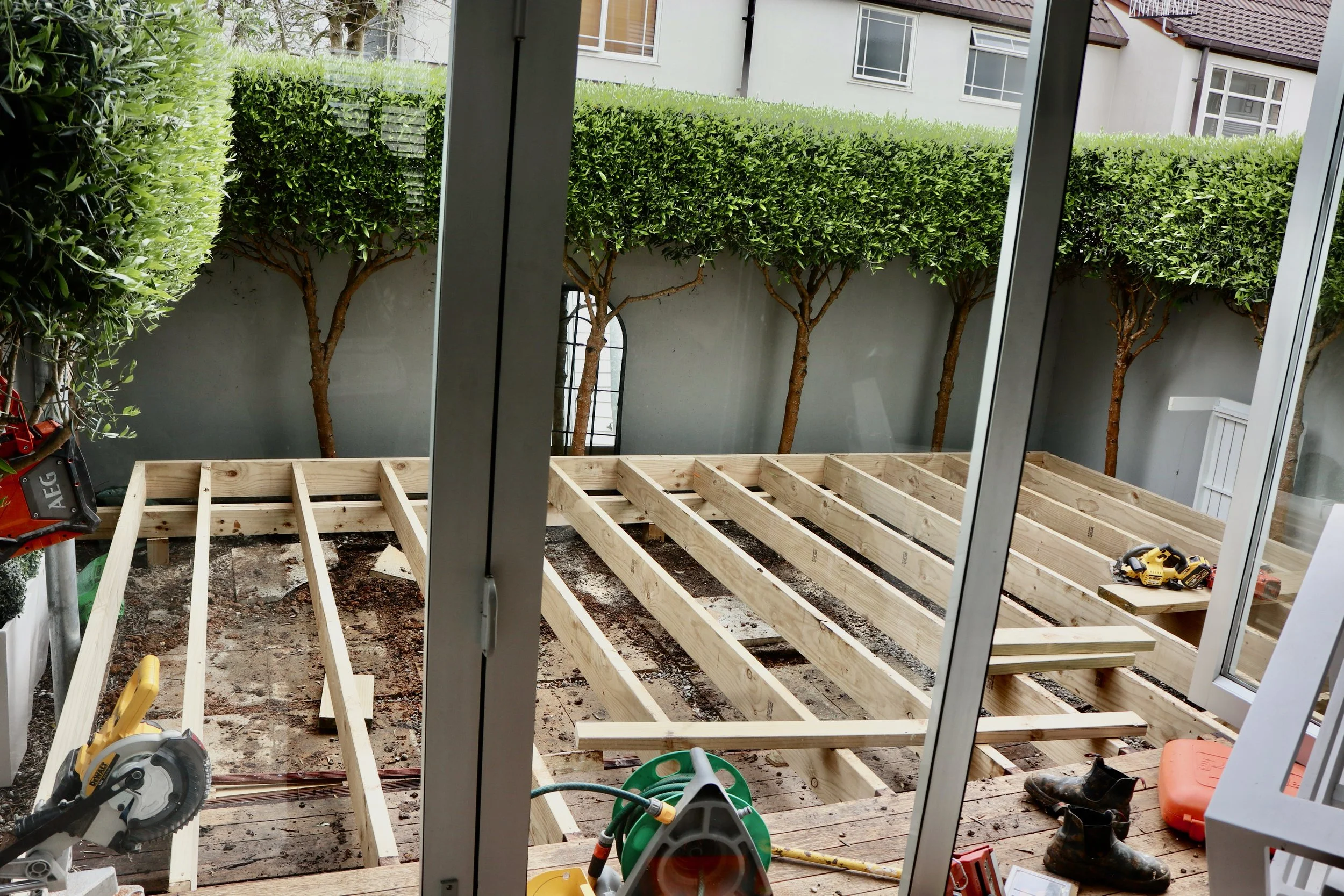 View of a backyard undergoing deck construction with wooden framing, tools, and equipment, seen through sliding glass doors.