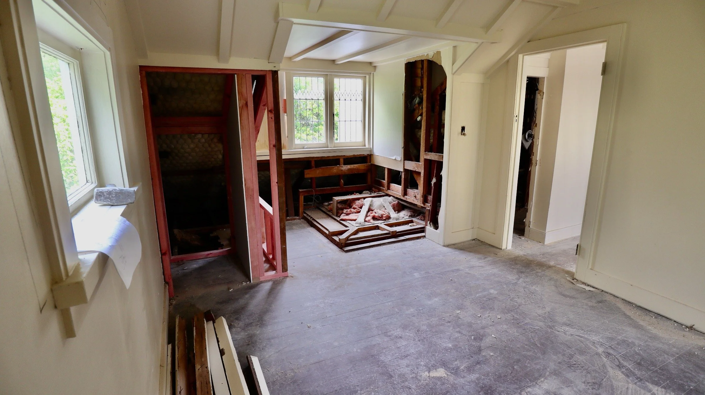 Room under renovation with exposed wooden framing, partially removed wall, and construction debris on the floor. Window with natural light.