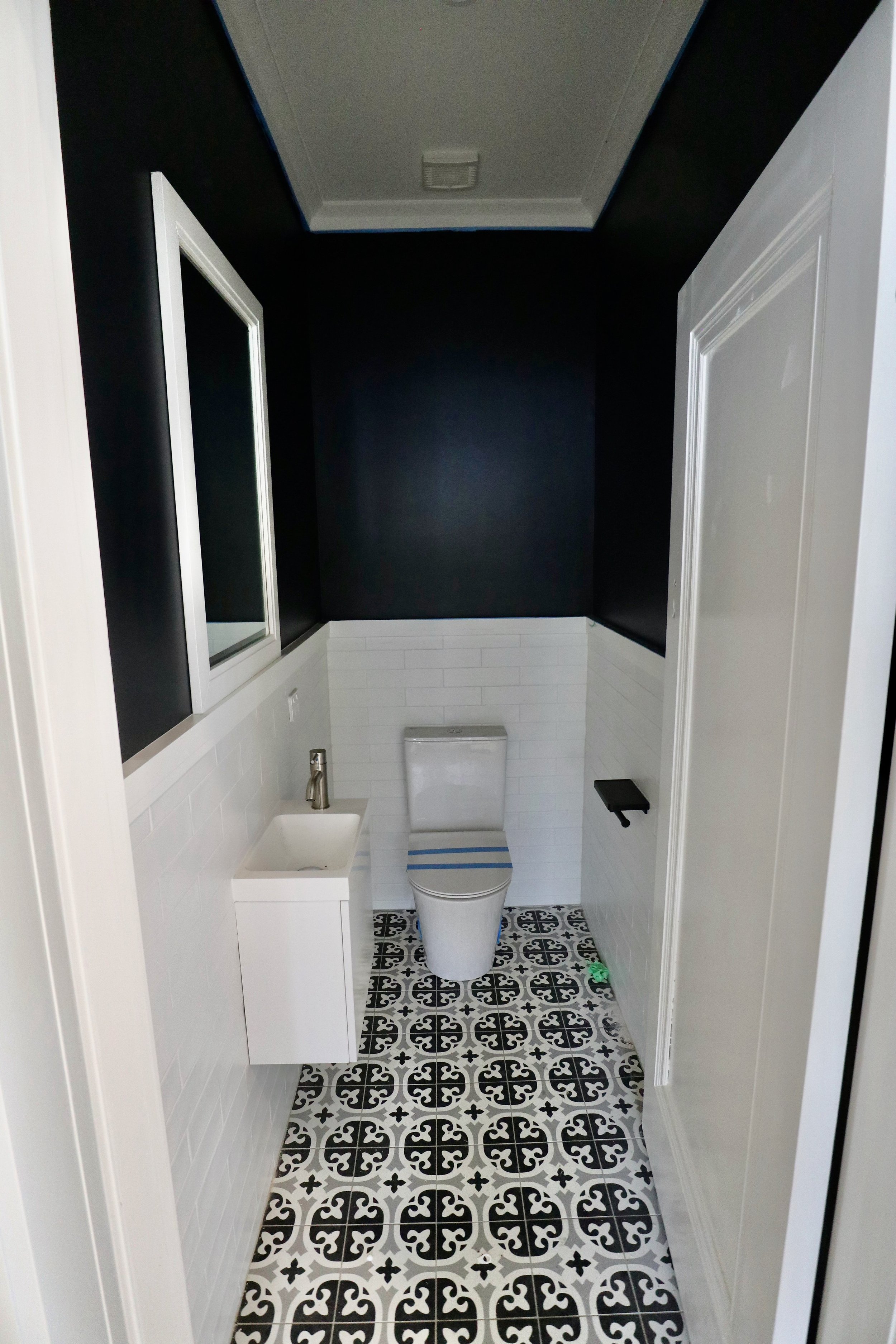 A small, modern bathroom with black and white decor, featuring a white toilet, a mini sink with a silver faucet, a black toilet paper holder, a mirrored medicine cabinet, and patterned black and white floor tiles.