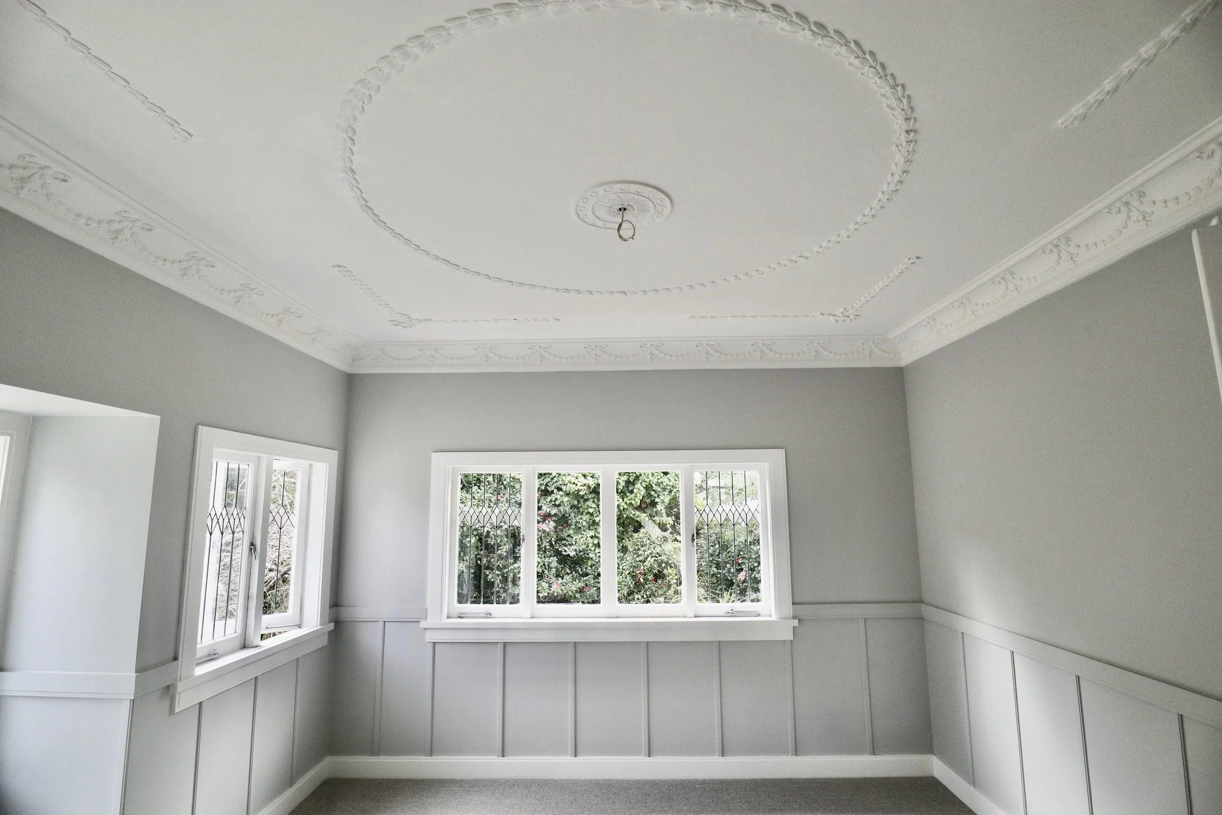 Empty room with light gray walls, decorative crown molding, and a ceiling medallion with a bulb socket, three-pane window with white trim, and view of greenery outside.