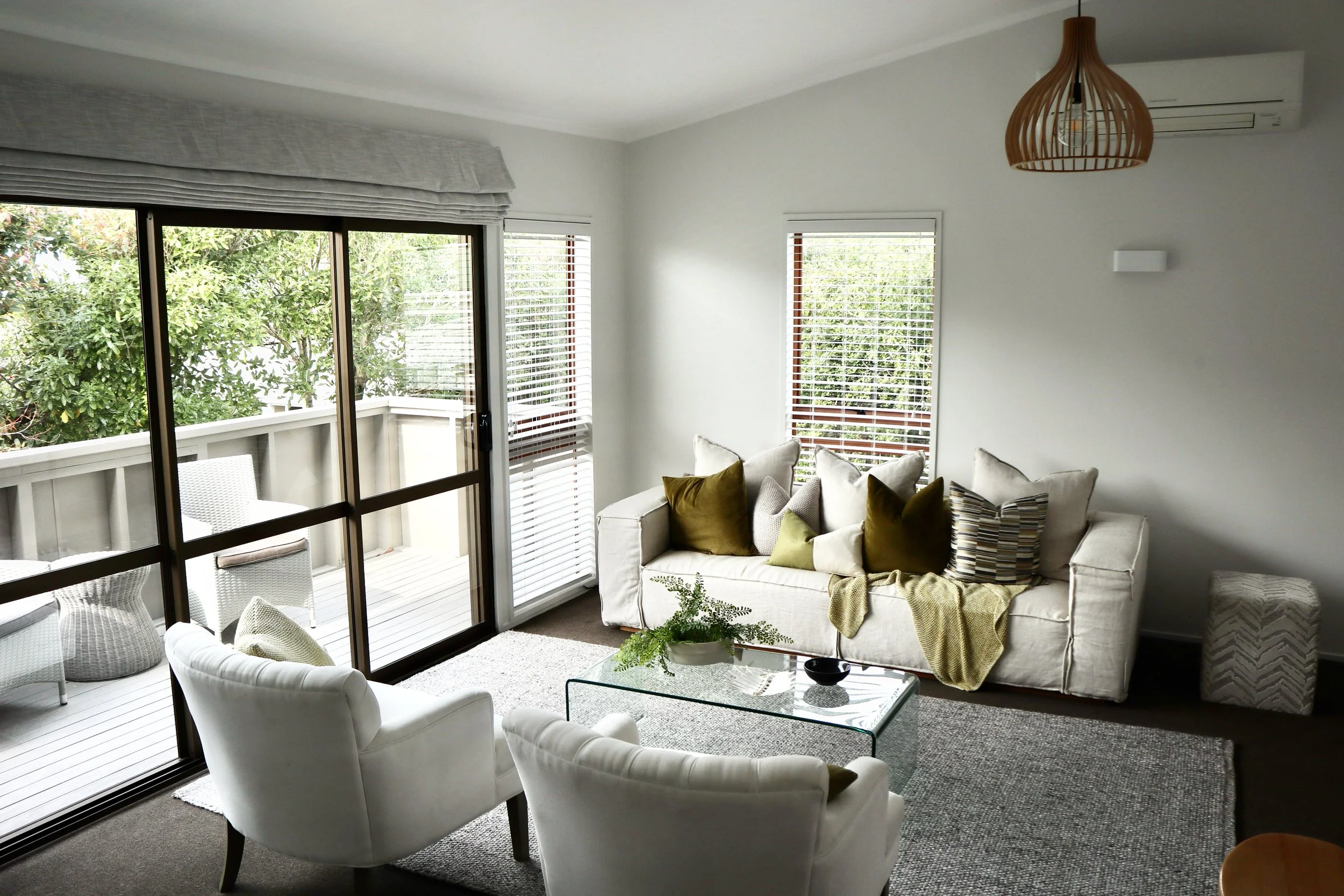 Living room with white sofa and armchairs, glass coffee table, sliding glass door leading to balcony, and large windows with blinds.
