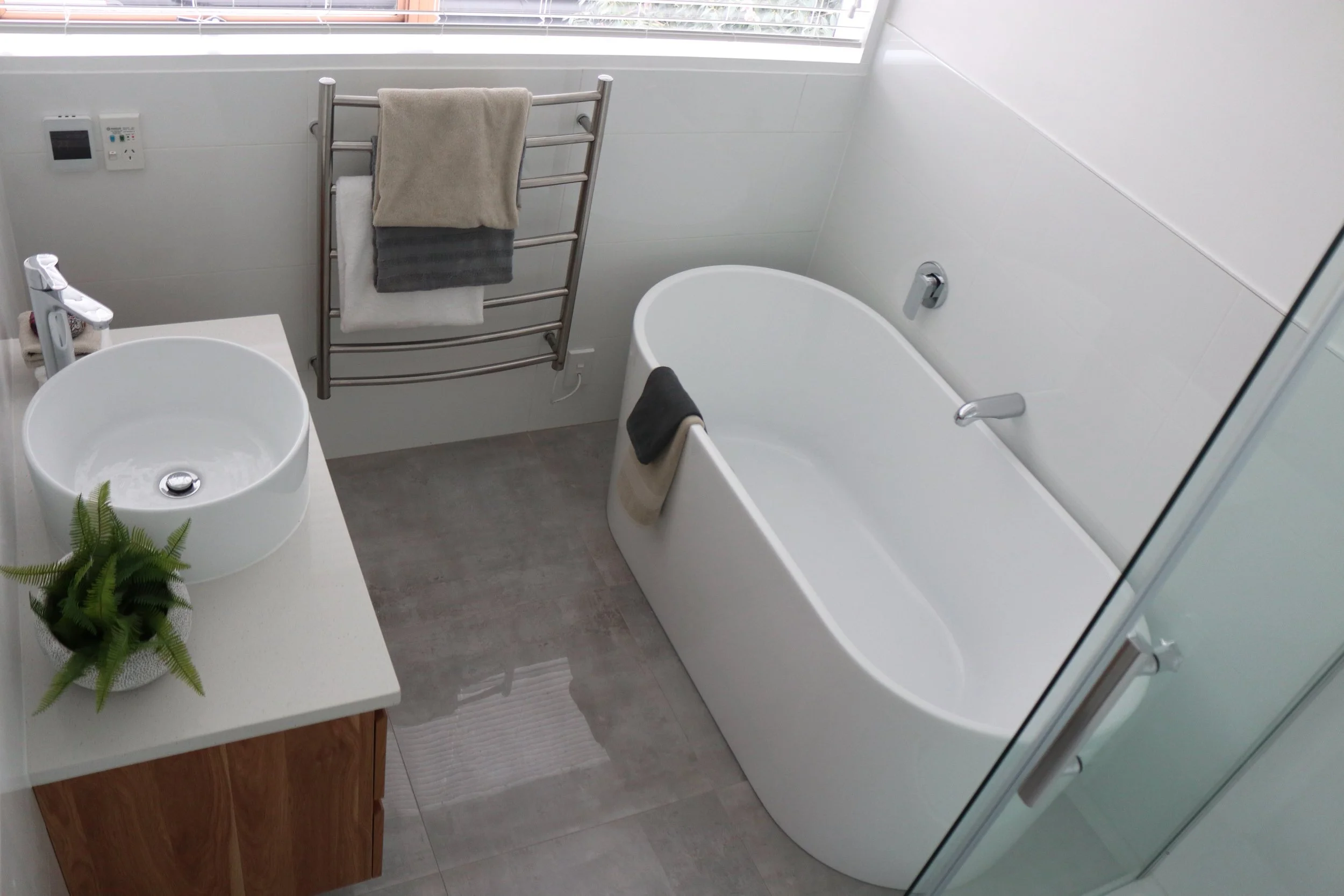 Modern bathroom with a freestanding bathtub, a round vessel sink on a wooden vanity, a towel rack with hanging towels, and a window with blinds.