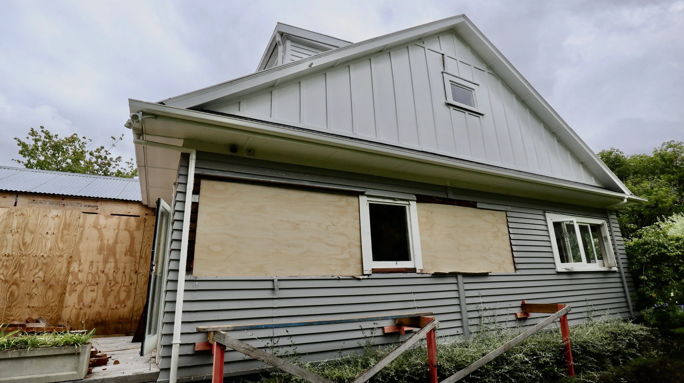 A house under renovation with a boarded-up window and an unfinished side wall, surrounded by some bushes and a wooden barrier in front.