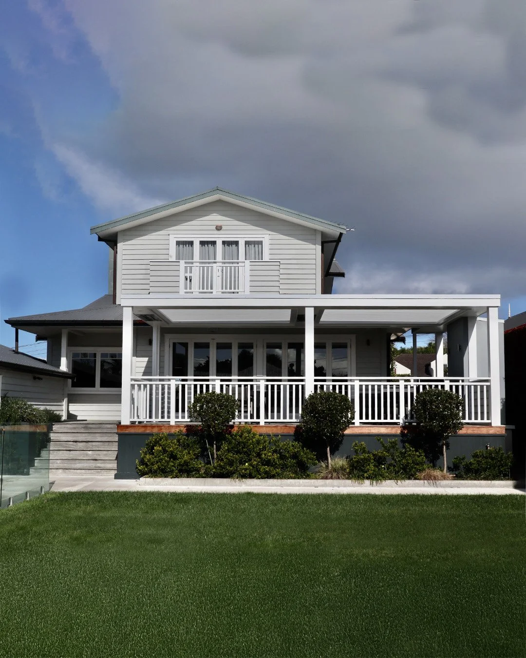 A two-story white house with a large front porch, white railing, and a small balcony on the second floor, surrounded by a well-maintained lawn and bushes under a cloudy sky.