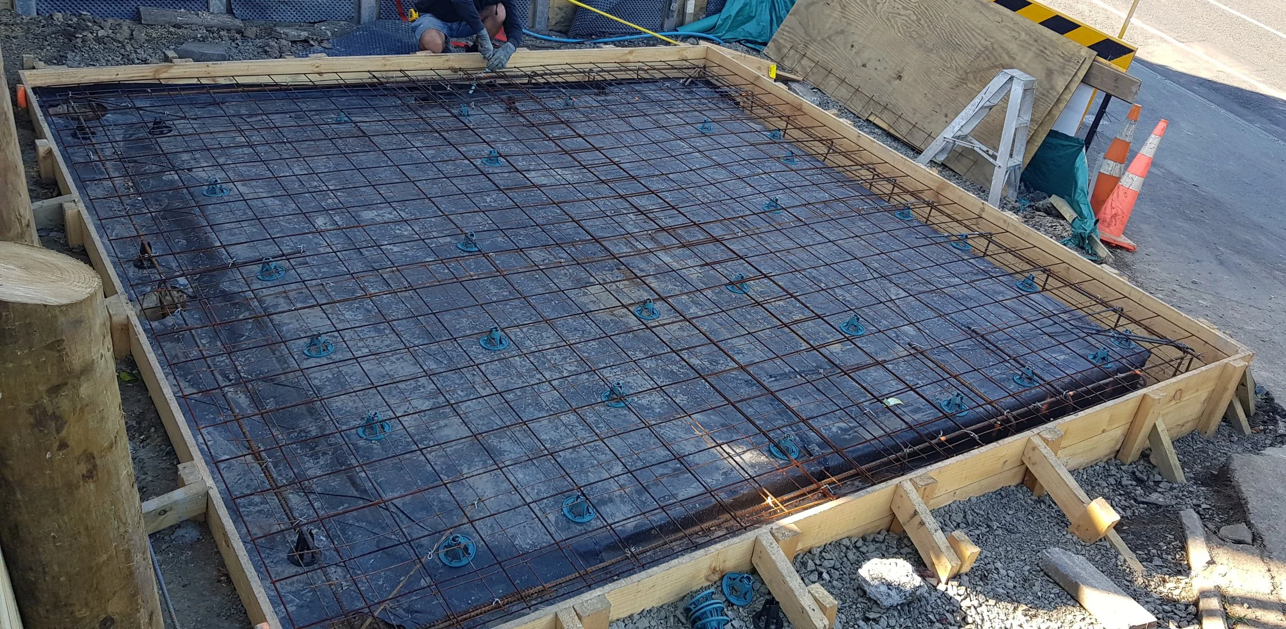 Construction site with rebar reinforcement framework for a concrete slab, surrounded by wooden forms and safety cones.