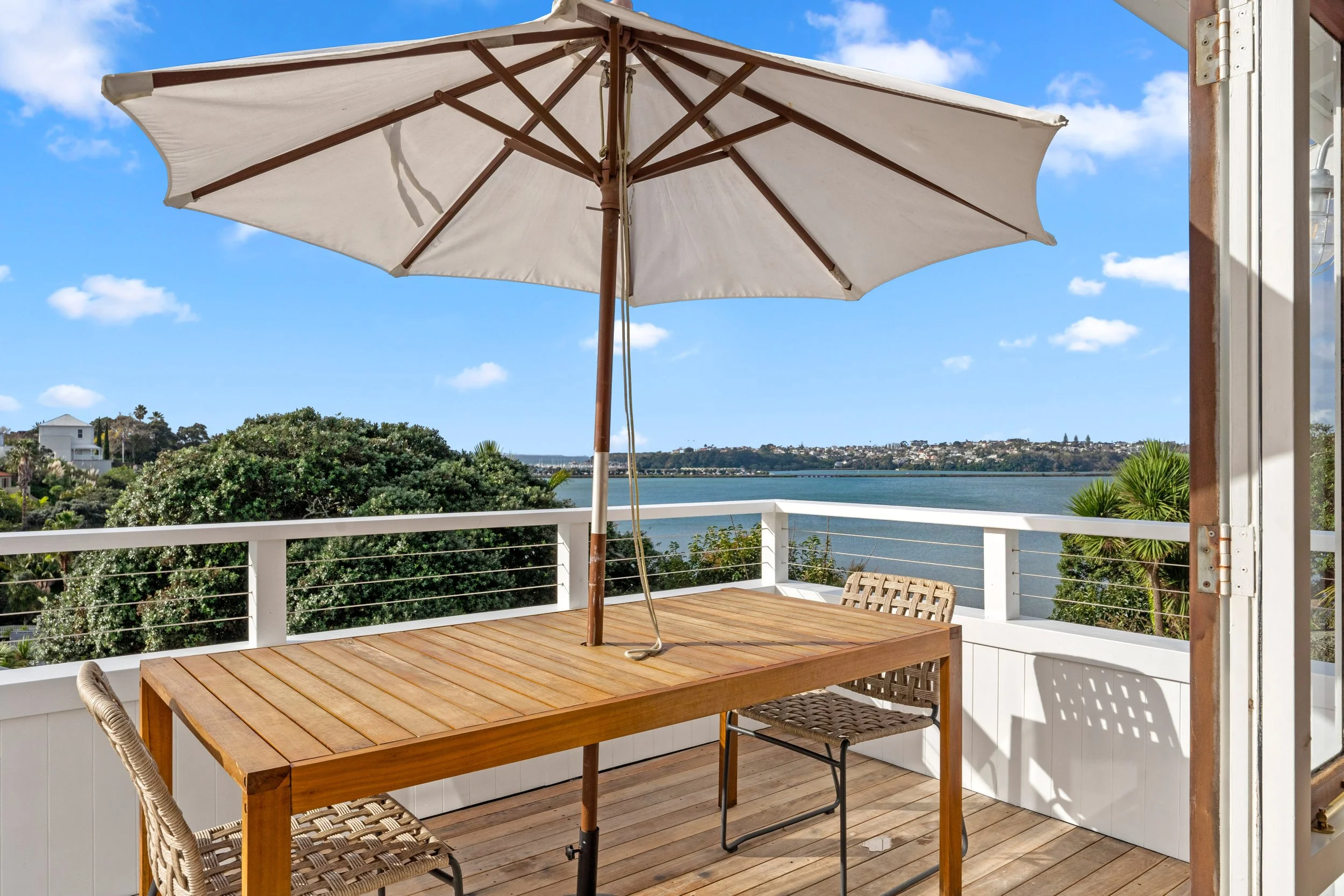A wooden deck with a table, two wicker chairs, and a large white patio umbrella, overlooking a body of water and green trees under a partly cloudy sky.