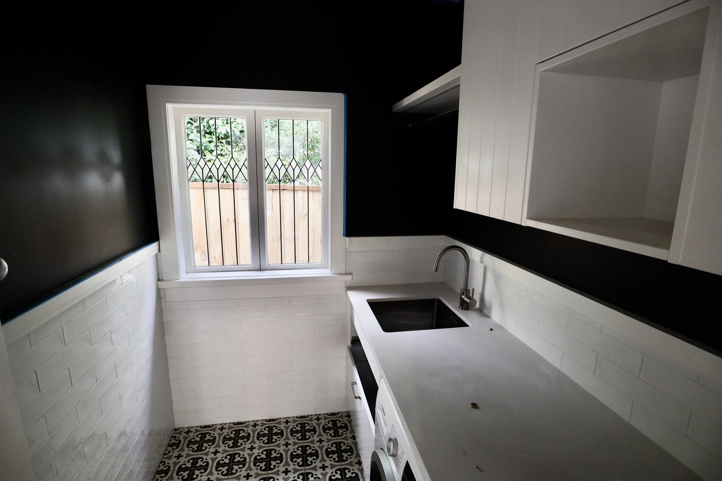 Small kitchen with black upper wall, white lower wall, white cabinets, black countertop, stainless steel sink, washing machine under countertop, window with security bars looking out to a wooden fence and trees, patterned floor tiles.