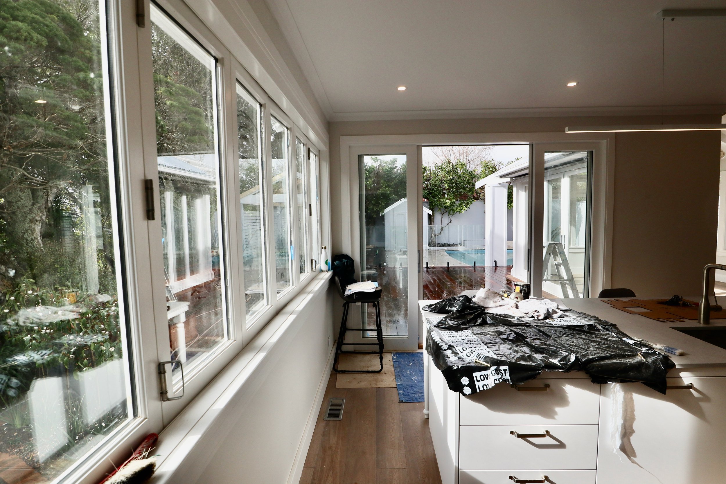 Interior view of a kitchen with a large window and sliding glass door leading to an outdoor patio and pool. The kitchen counter has a black plastic bag and miscellaneous items on it. There is a small stool with clothing or fabric on top near the slid