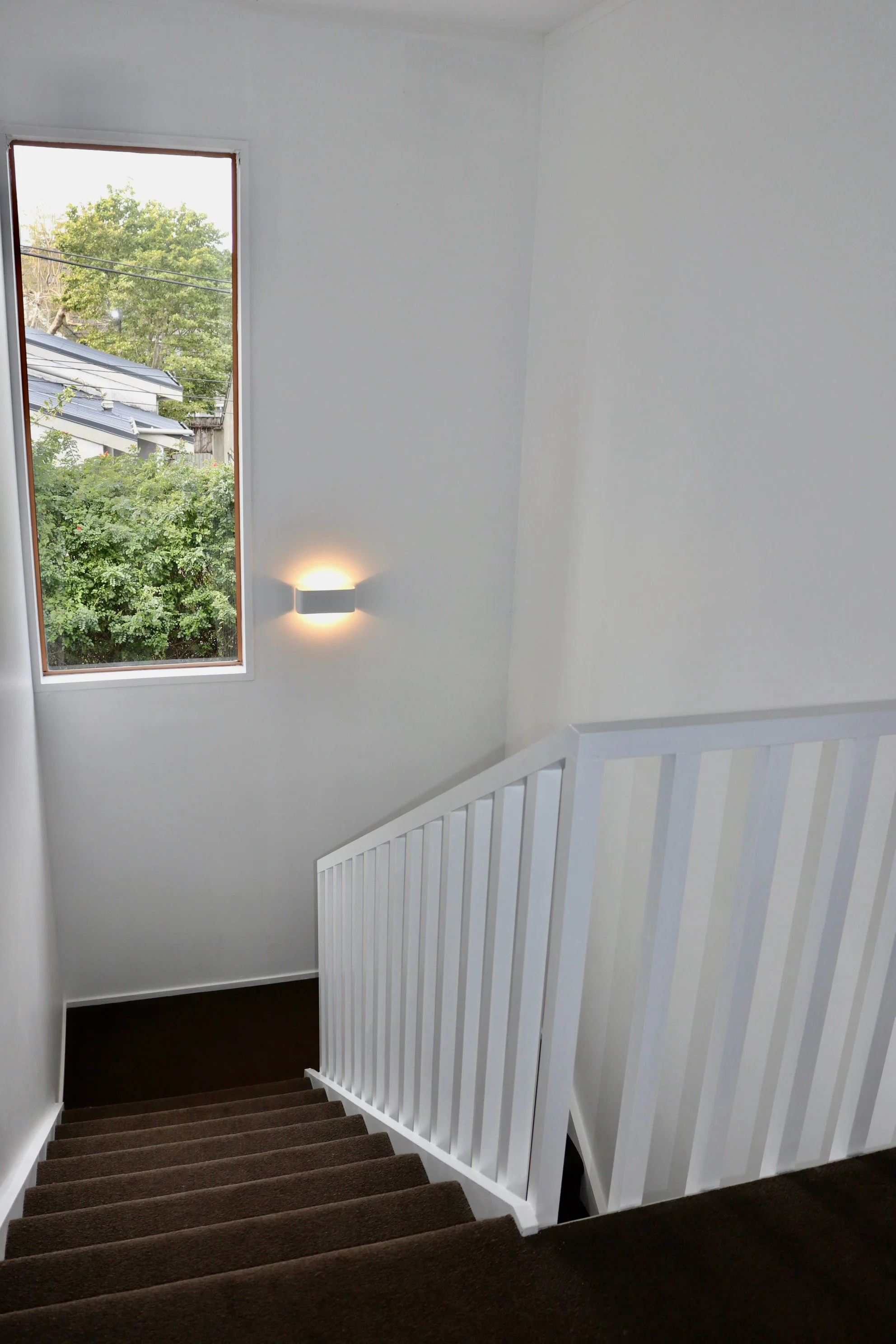 An interior staircase area with brown carpeted stairs, white railing, a wall-mounted light fixture, and a tall window showing trees and neighboring buildings.
