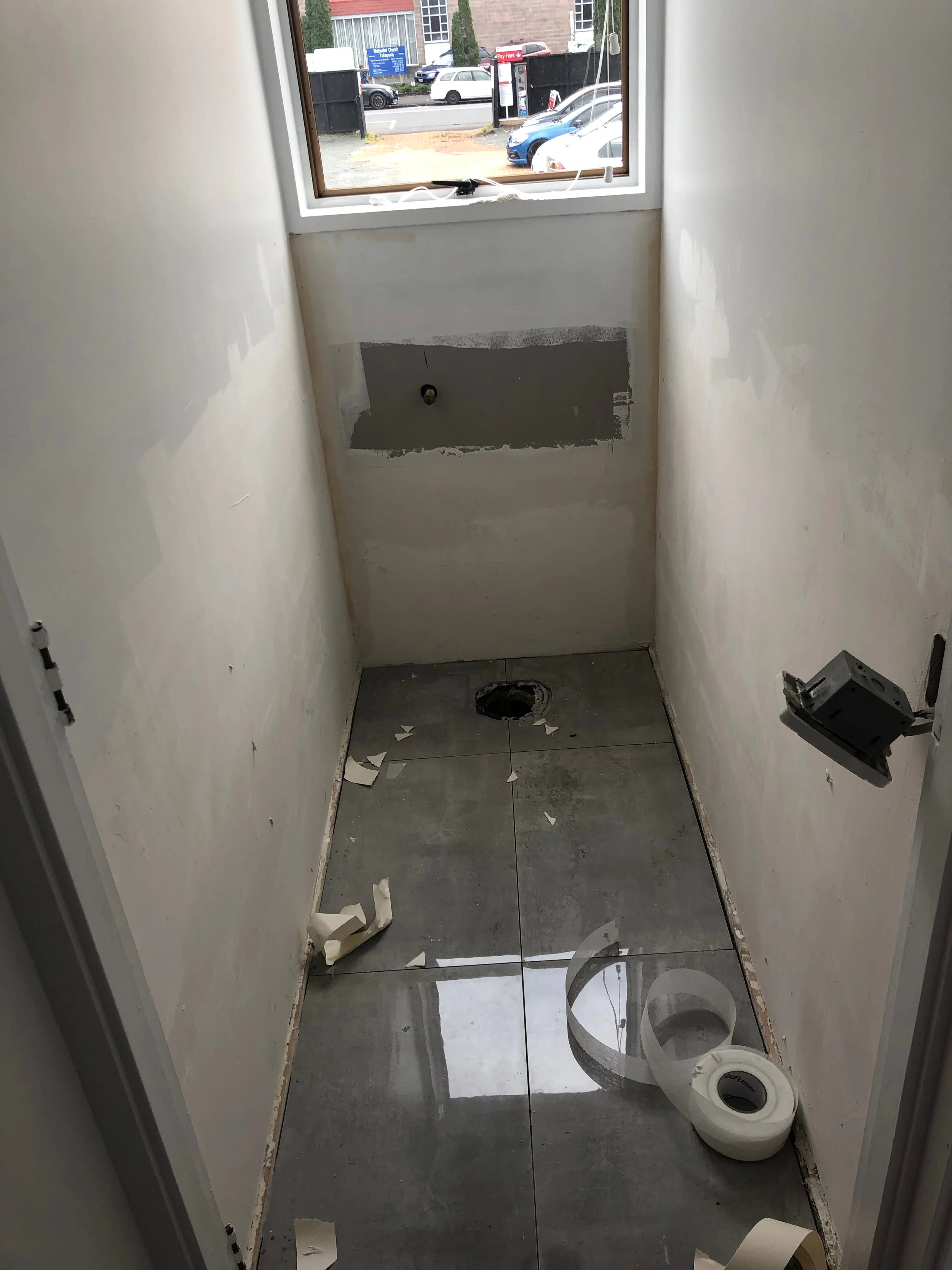 A small room under renovation with tiled floor, a hole in the floor, an unfinished wall with a gray patch, a small window looking outside, and construction debris including rolls of tape and torn paper scattered around.