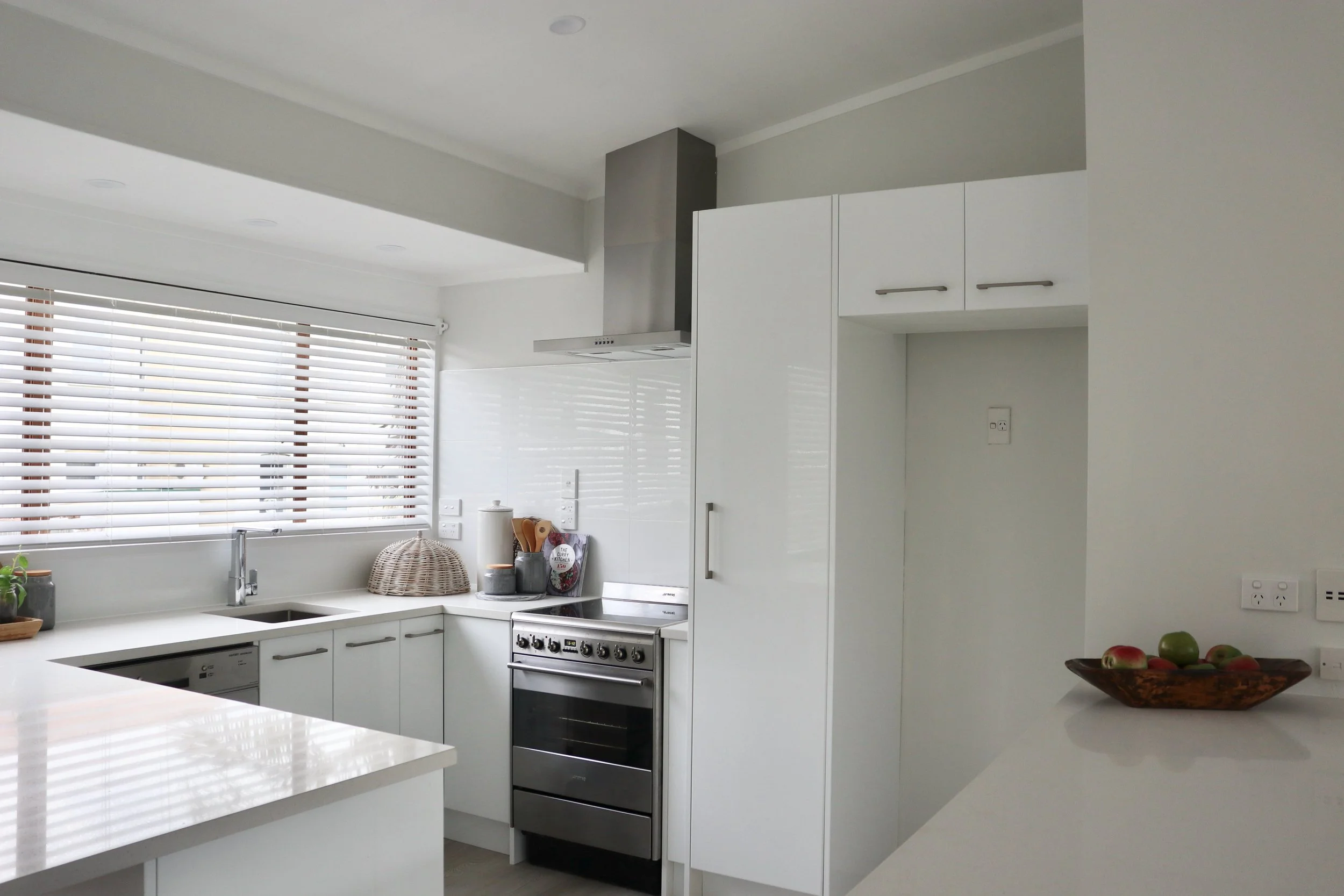 Modern white kitchen with a window with blinds, stainless steel stove, white cabinets, and a bowl of apples on a countertop.