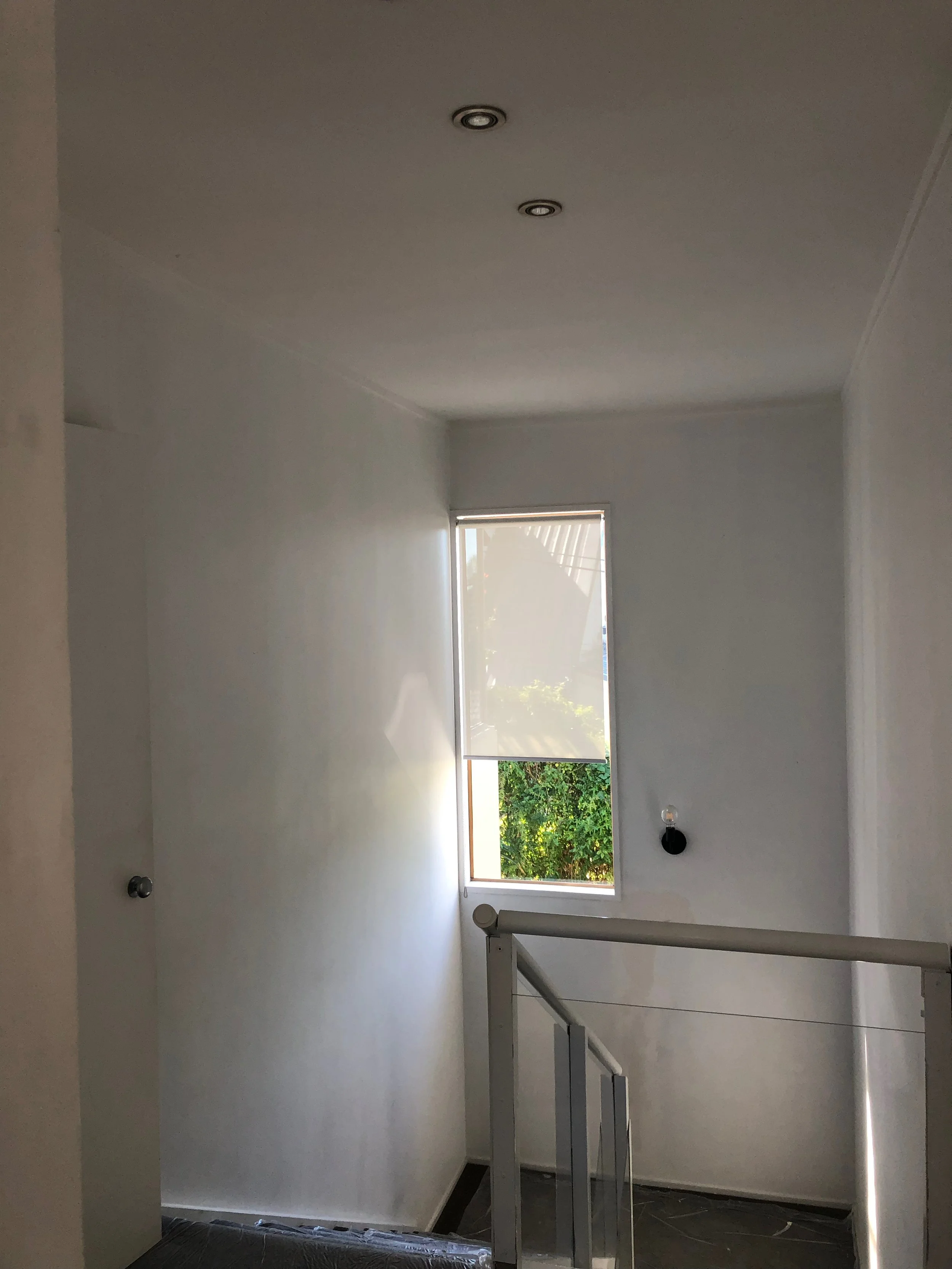 Empty room with white walls, a window with blinds, two ceiling lights, and a staircase railing.
