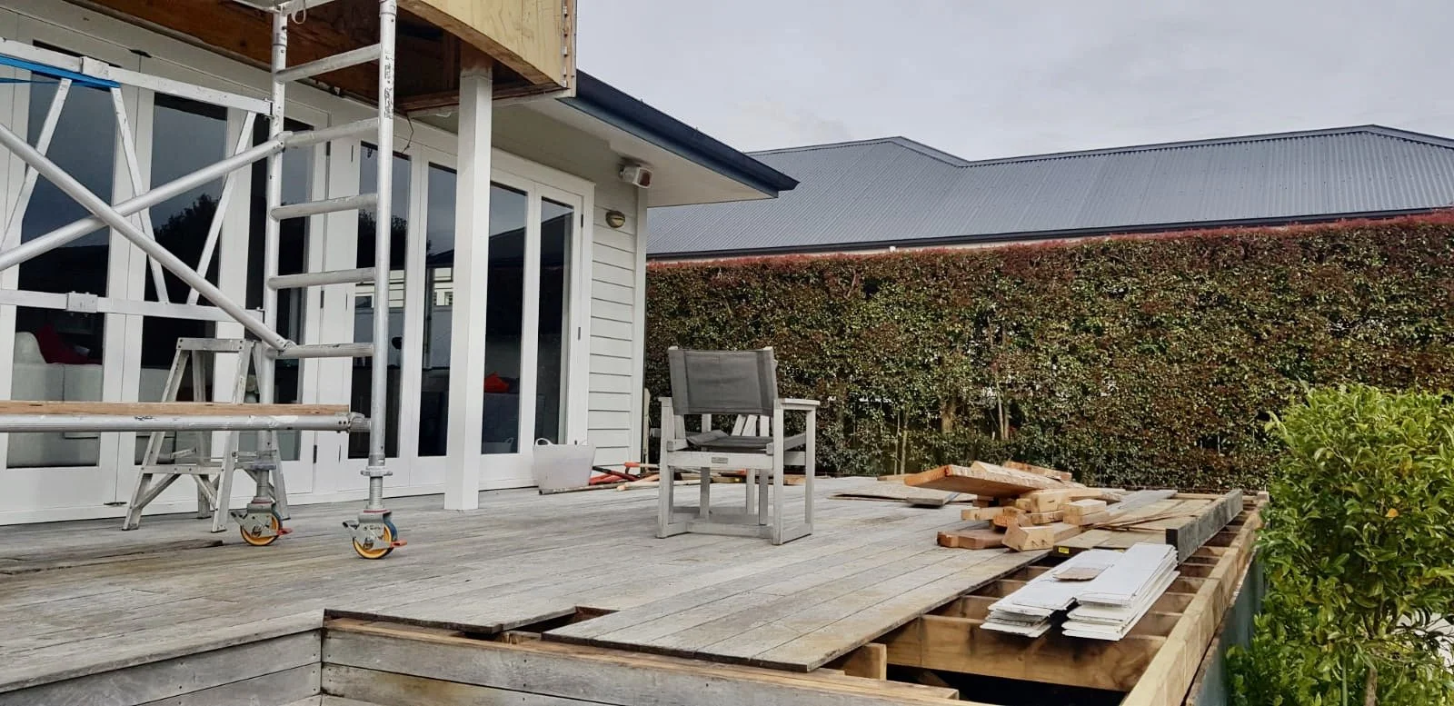 A deck under construction with wooden planks, scaffolding, an empty chair, and construction materials. A hedge and part of the house with sliding glass doors are visible in the background.