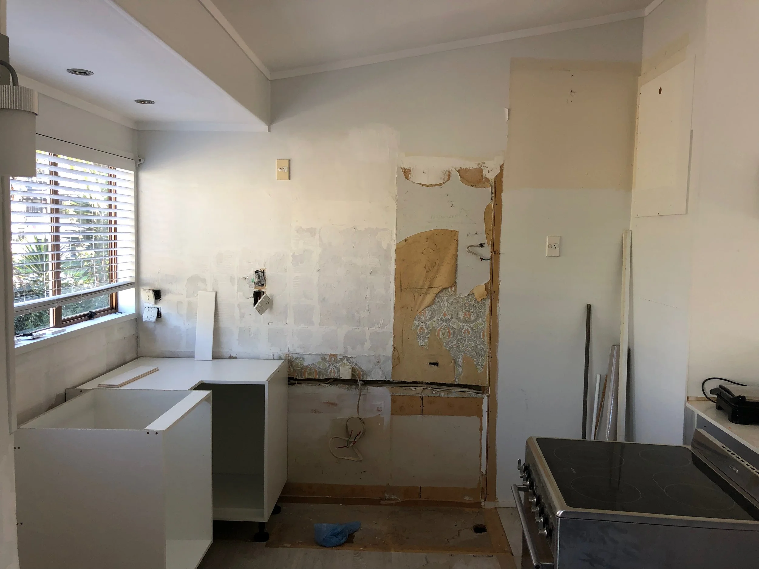 A kitchen undergoing renovation with exposed wall area, a stove on the right, and a white cabinet on the left under a window with blinds.